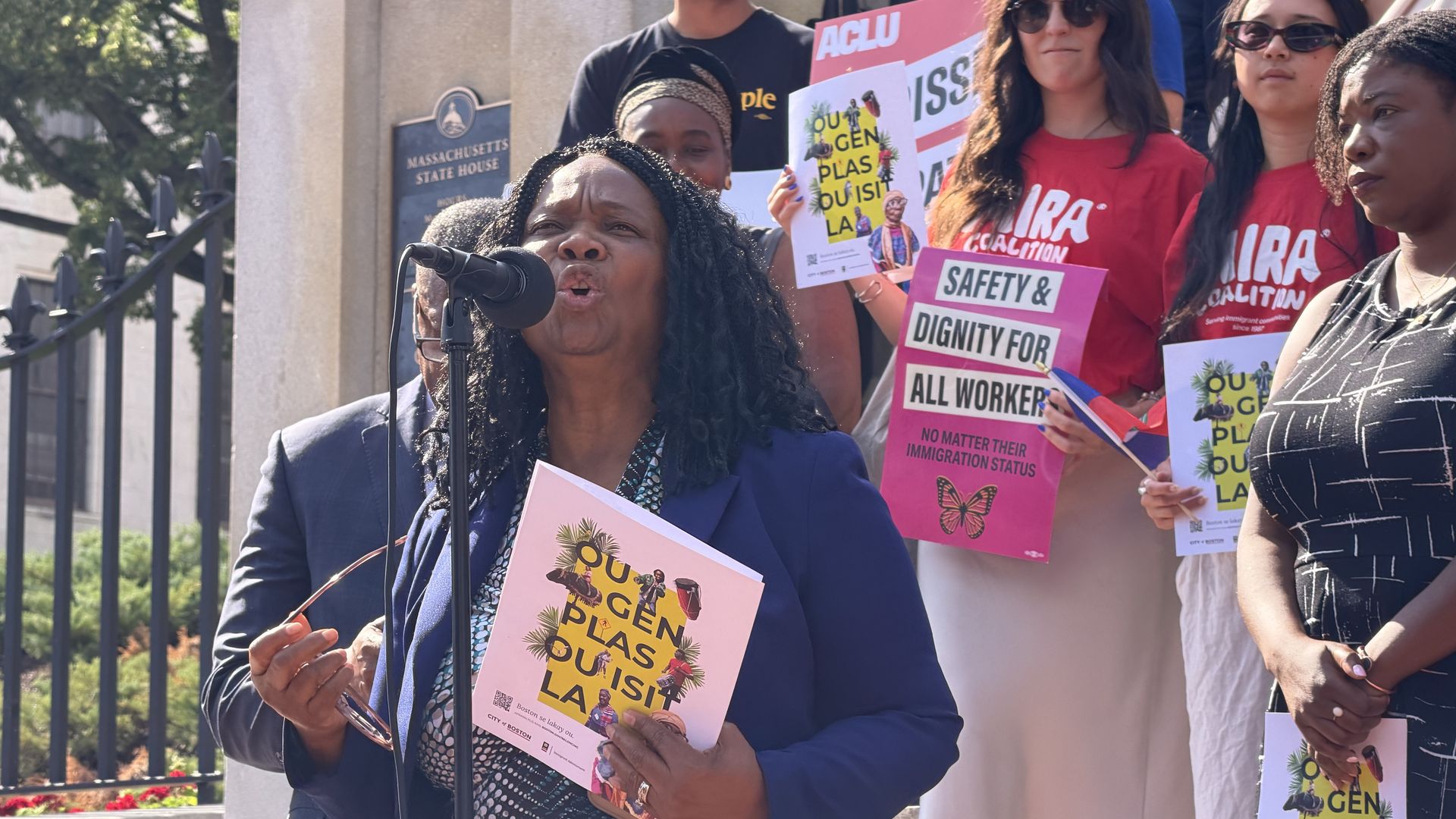 Geralde Gabeau, IFSI-USA's founder and executive director, speaks in front of the Massachusetts State House's front steps as Boston City Councilor Ruthzee Louijeune and other supporters stand behind her.