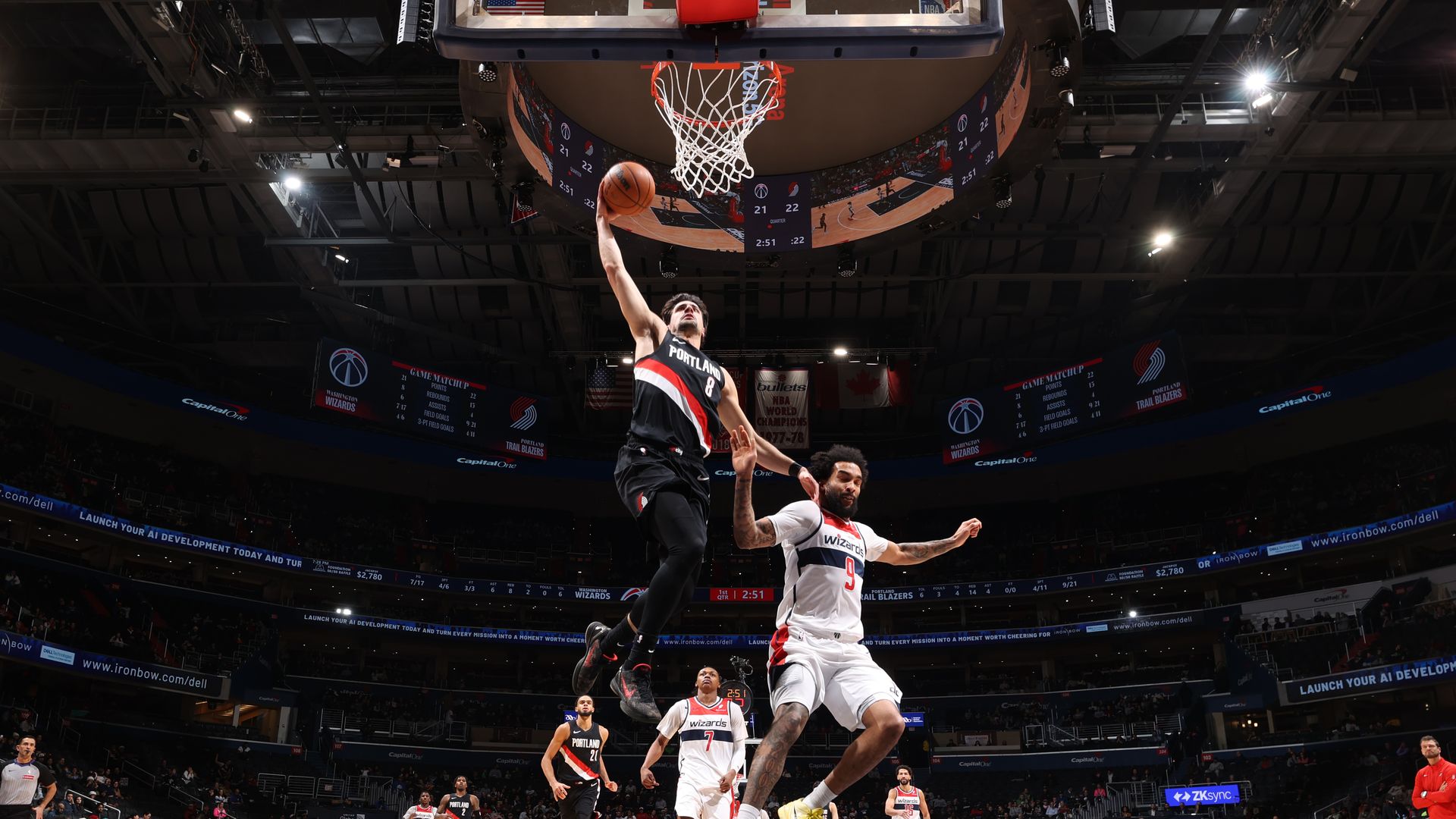 Basketball player from Portland Trail Blazers in black jersey jumps for a layup as a Washington Wizards player in white jersey with yellow shoes defends under the hoop during a game.
