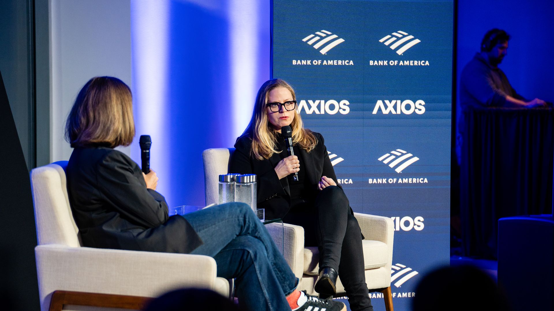 Two women in black outfits sit in beige chairs on a stage, holding microphones in conversation, with a blue backdrop showing "Bank of America" and "AXIOS" logos, a man in headphones stands nearby.