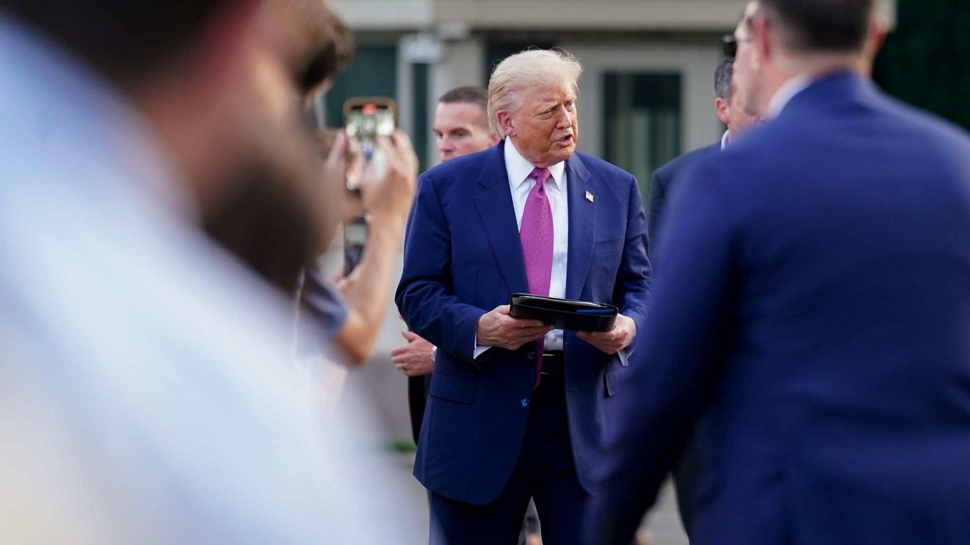 President Trump, wearing a blue suit and holding a folder while surrounded by security, aides and others outside the White House.