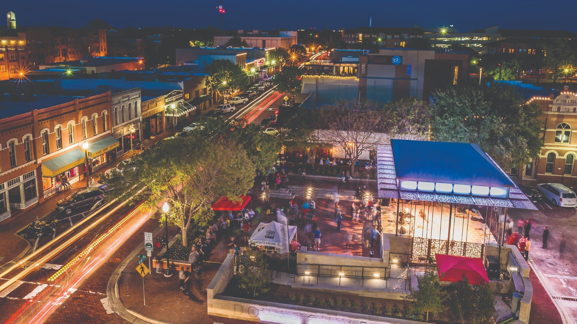 Aerial night view of a busy downtown street with brick buildings, string lights, and trees. Long-exposure car streaks flow along the avenue toward a plaza with people and a blue-roofed pavilion.