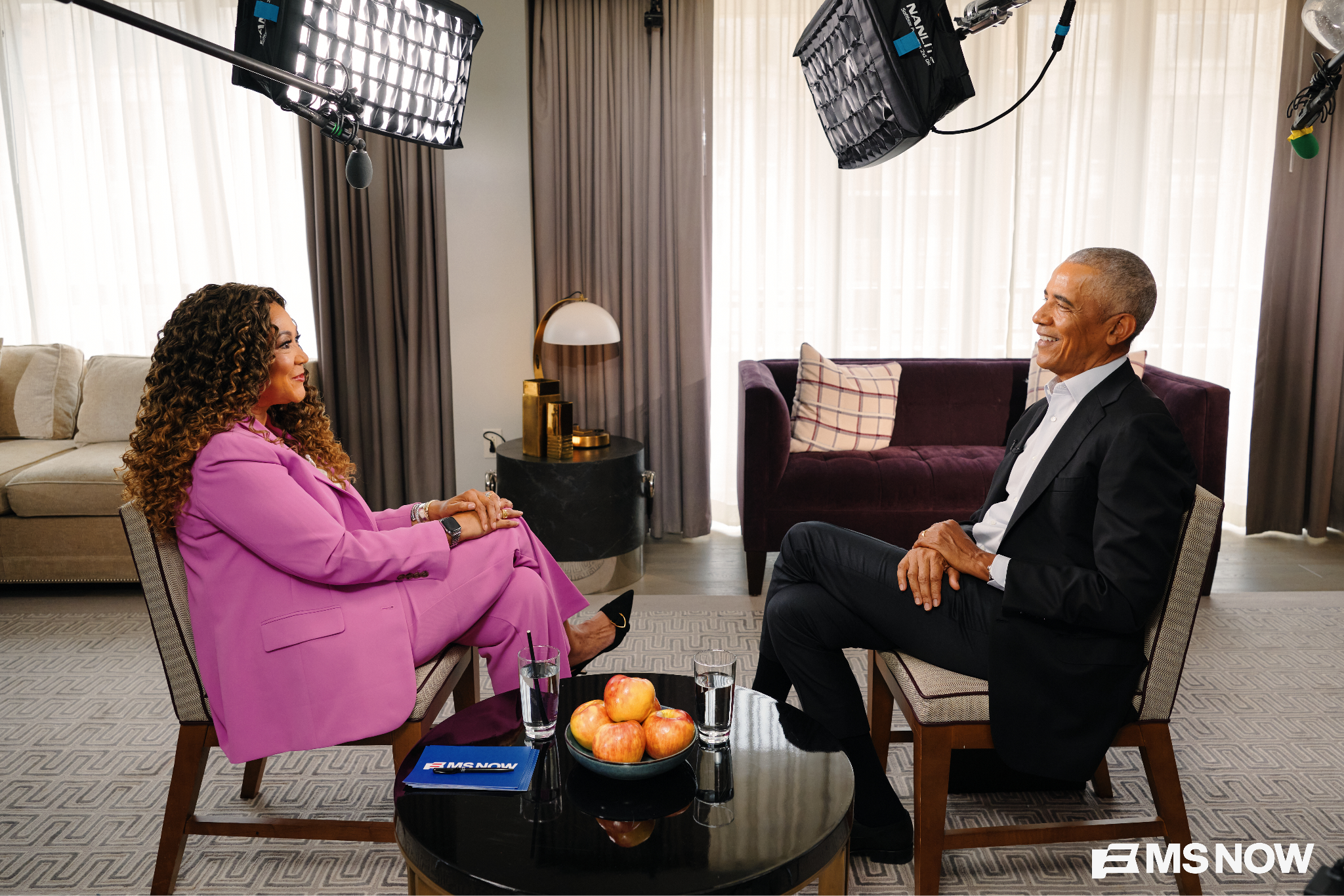 A woman in a pink suit and former President Obama, wearing a black suit and a white collared shirt, sit in a living-room studio sit facing each other. A glass coffee table with apples and water glasses sits between them; studio lights hang overhead.