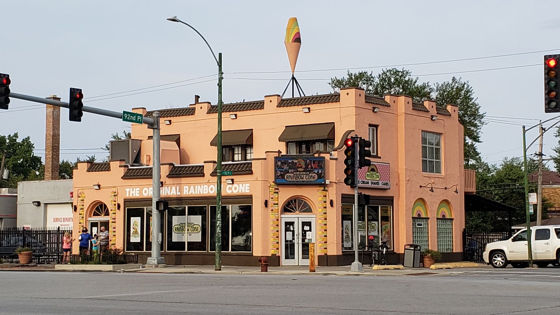 Peach-colored building at street corner with signs reading "The Original Rainbow Cone". Colorful ice cream cone sculpture on roof. Traffic lights show red. Four people standing outside.
