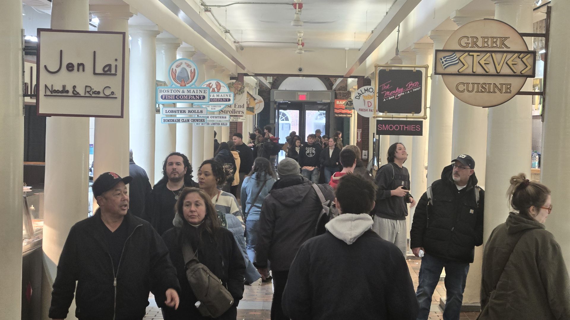 Busy indoor market corridor with white columns and hanging food signs. Left sign reads "Jen Lai Noodles & Rice Co."; right round sign advertises Greek cuisine. Crowded with shoppers in coats.