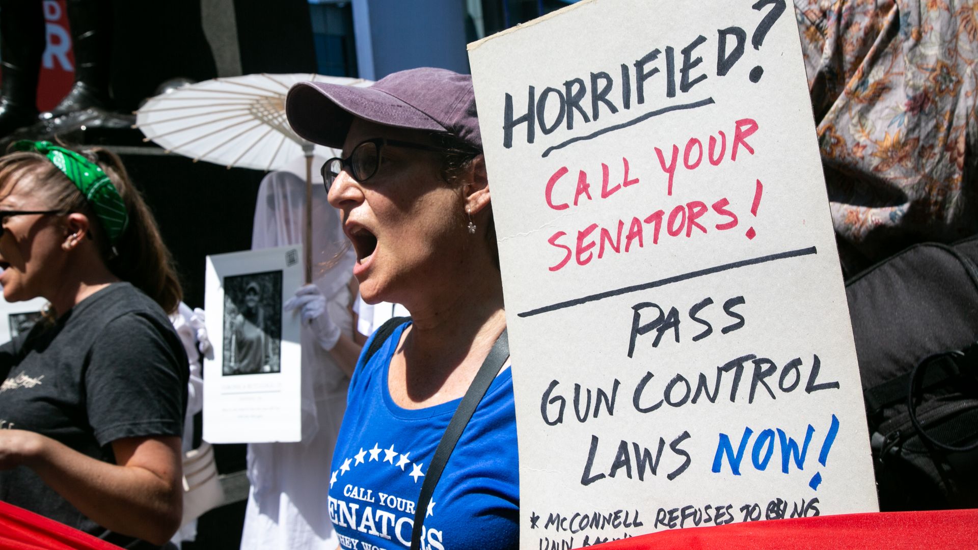 Picture of a woman at a protest holding a sign that says, "horrified? call your senators! pass gun control laws now!"