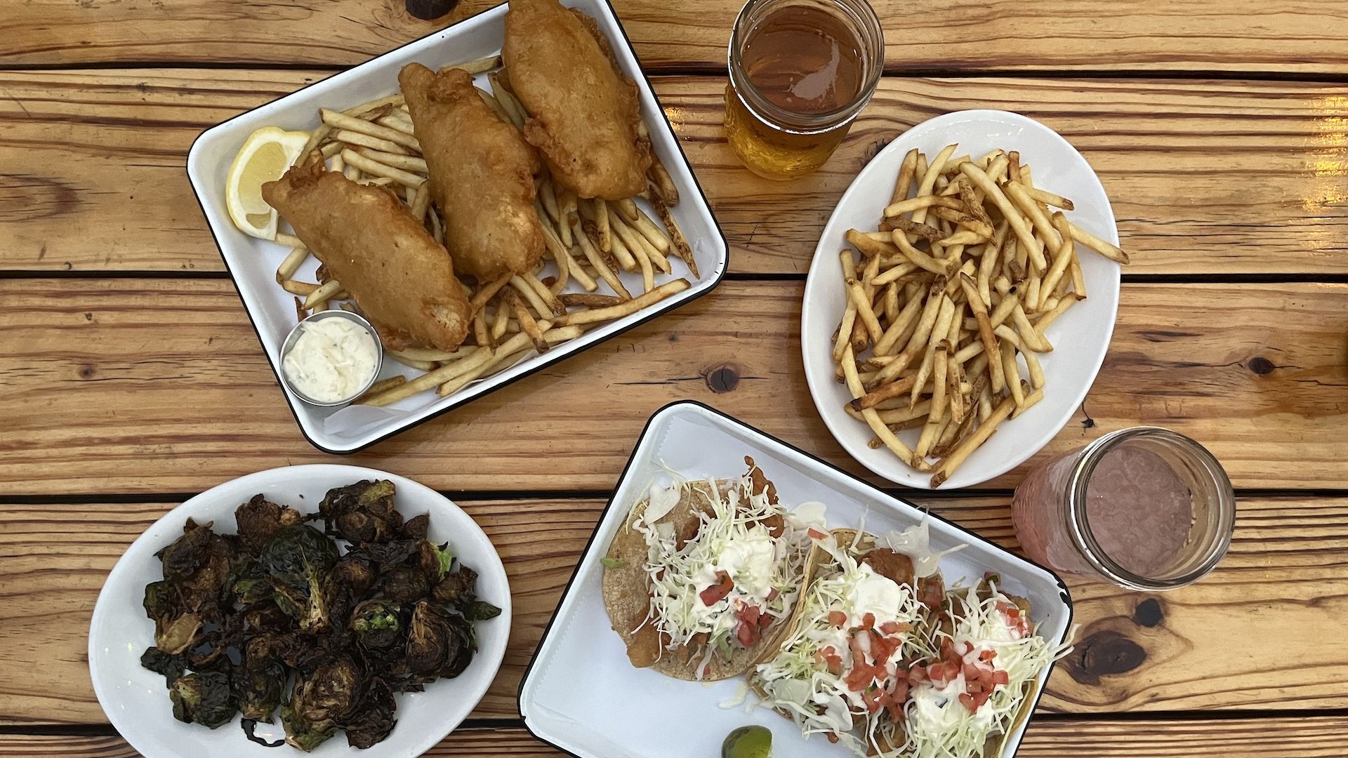 Plates of fish and chips, brussels sprouts, baja fish tacos and fries with two drink on a picnic table.