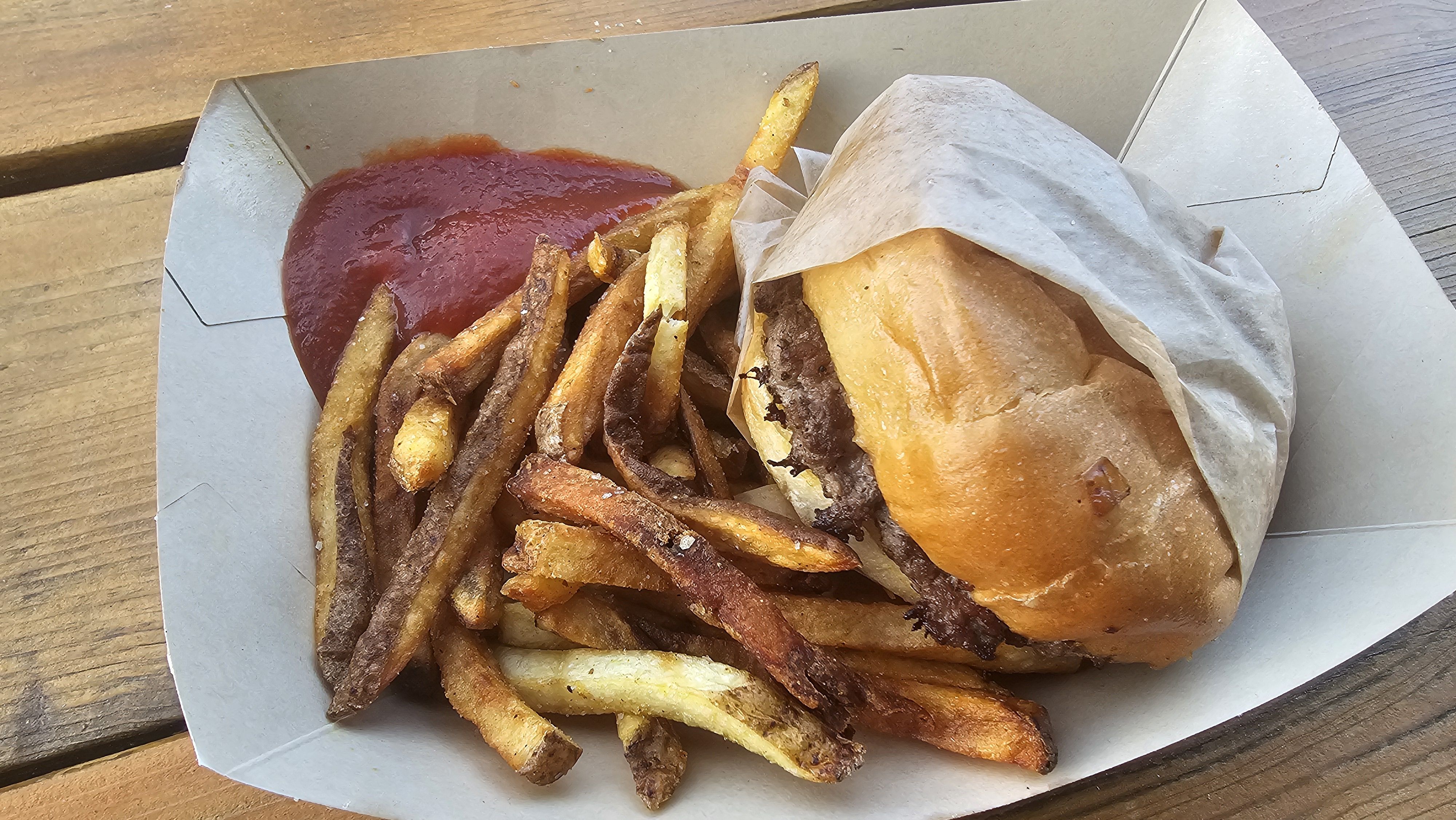 Open takeout container on a wooden table, featuring a wrapped hamburger and a mound of french fries with a side of red ketchup.