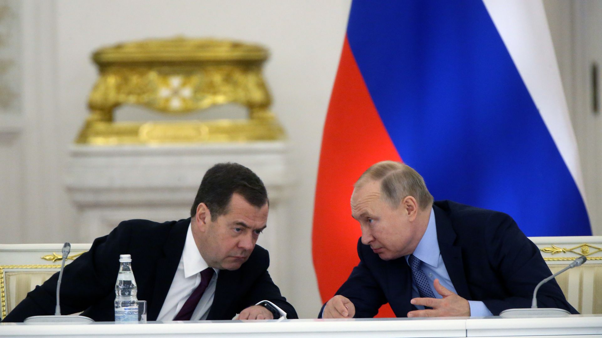 Two men in dark suits leaning towards each other whispering at a white table with microphones and a water bottle. Russian flag and ornate gold decoration in background.