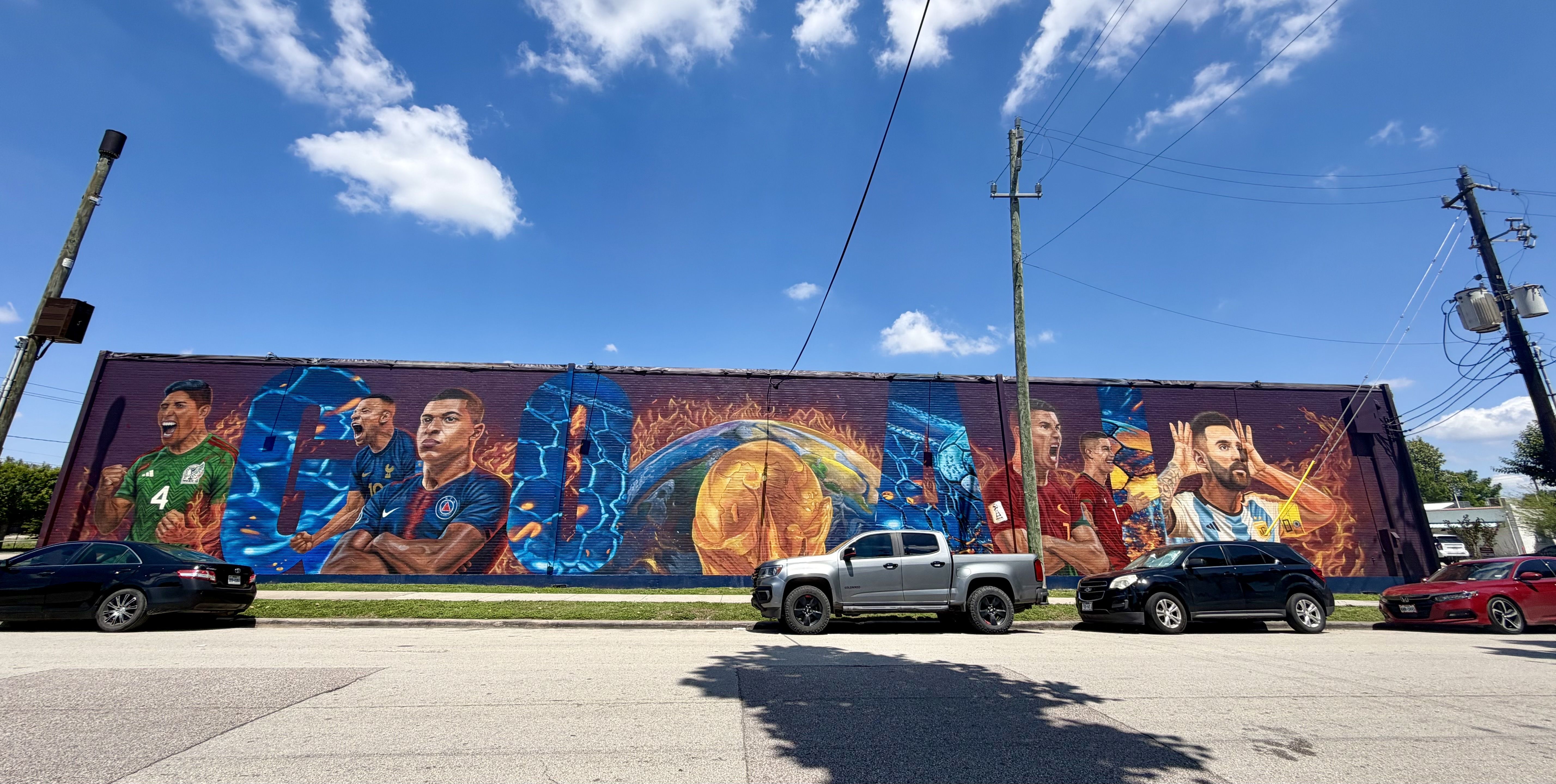 Wide mural on a brick wall depicting stylized soccer players in green, blue, and red jerseys amid flames and a globe motif; power poles, blue sky with clouds, and parked cars in front.