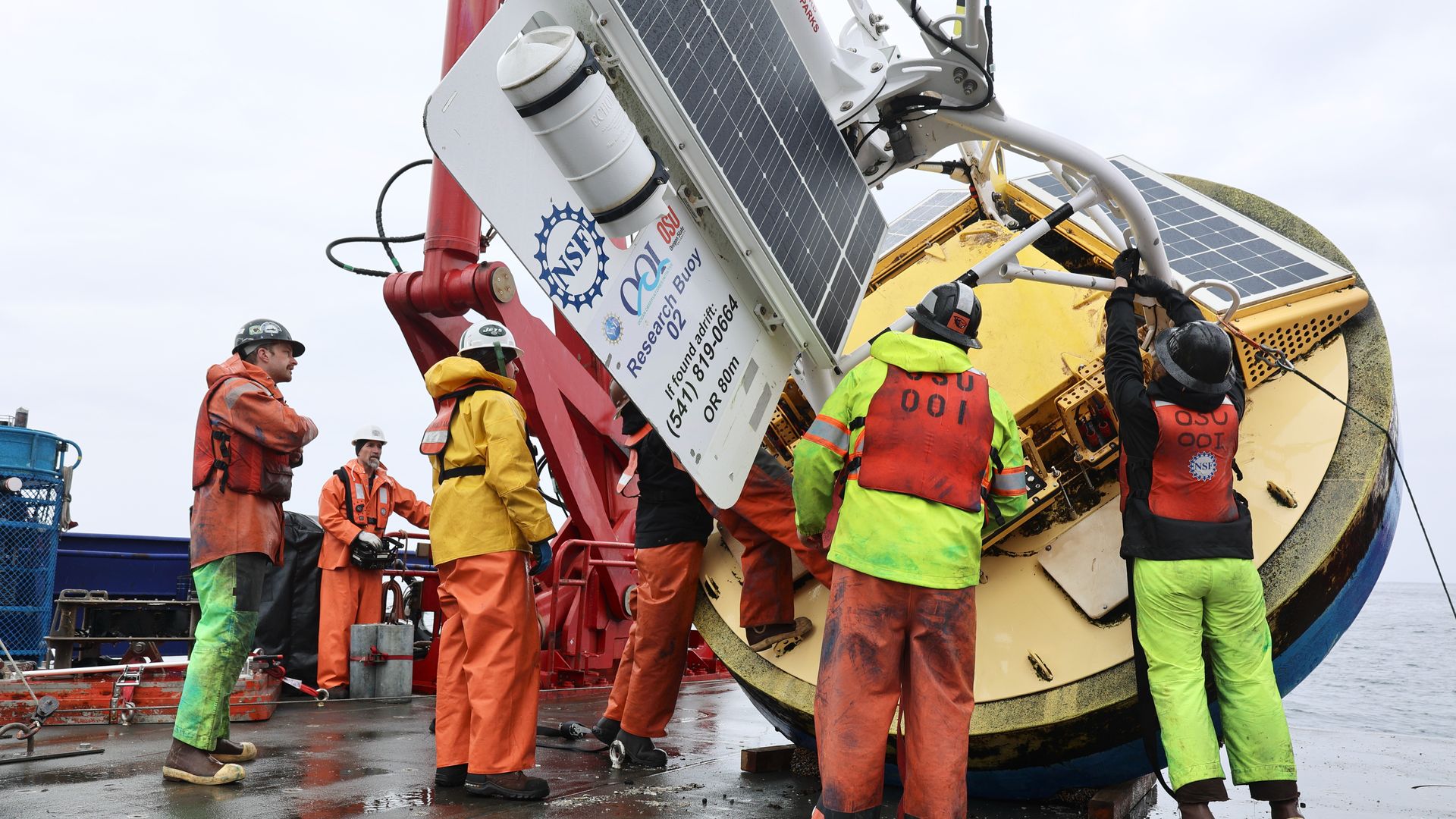 Workers in orange and high-visibility gear inspect a large yellow circular machine with a white frame and solar panels, as a red crane arm looms nearby on a wet deck.