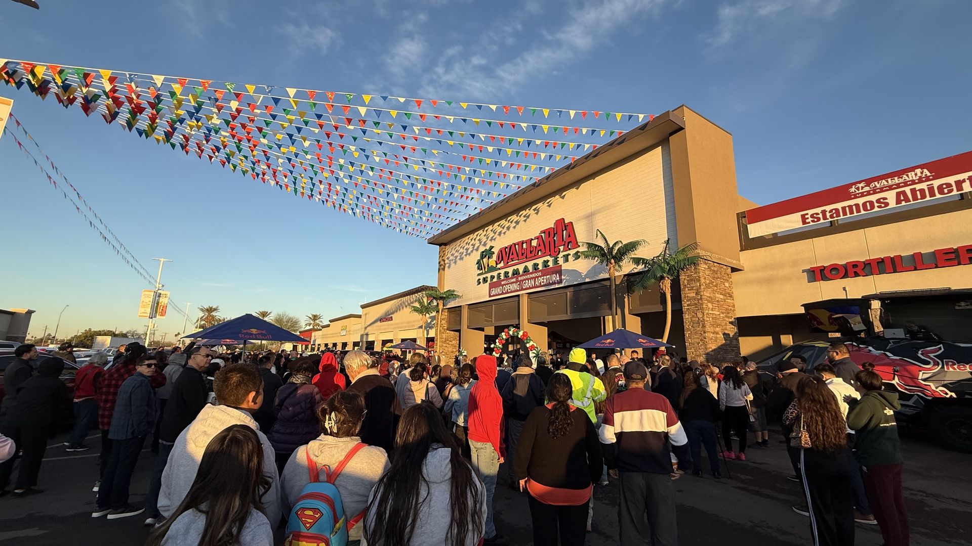 Crowd gathered outside Vallarta Supermarket at golden hour with colorful pennant flags overhead and festive decorations for grand opening event.