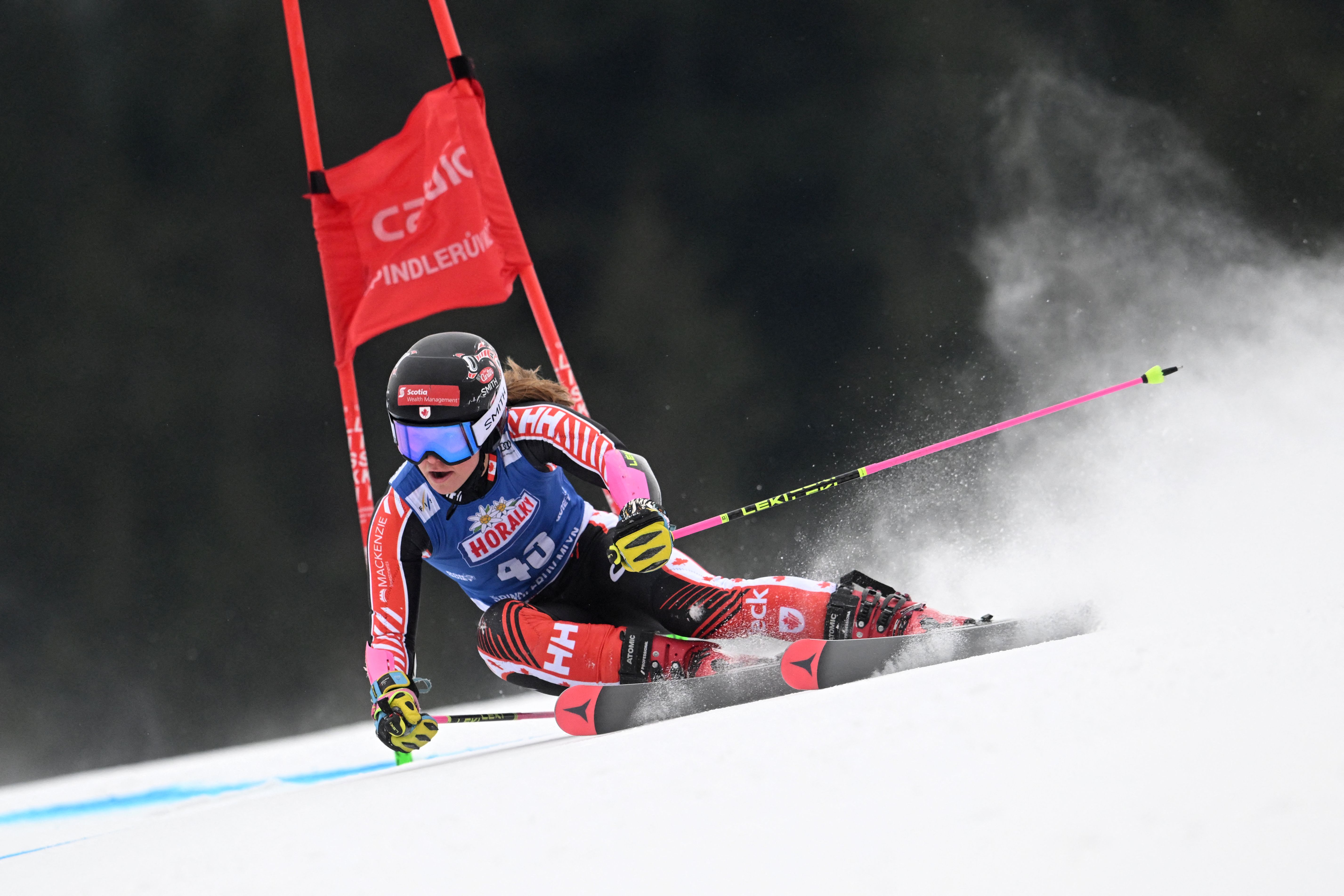 Skier in red and black suit with blue bib, wearing a black helmet and goggles, making a sharp turn on a snowy slope near a red gate with white text, kicking up snow spray.