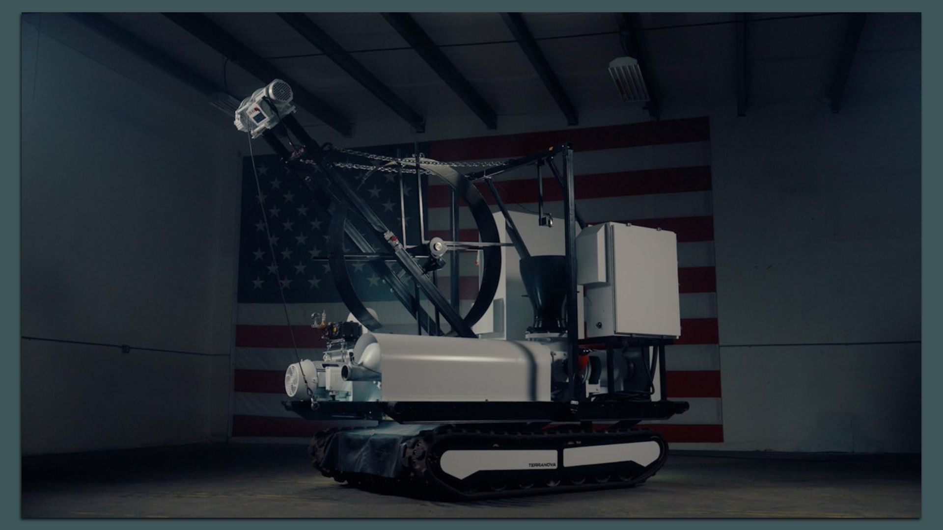 Gray and black robotic machine, sitting on tank-like treads, inside a dimly lit room with an American flag on the back wall.