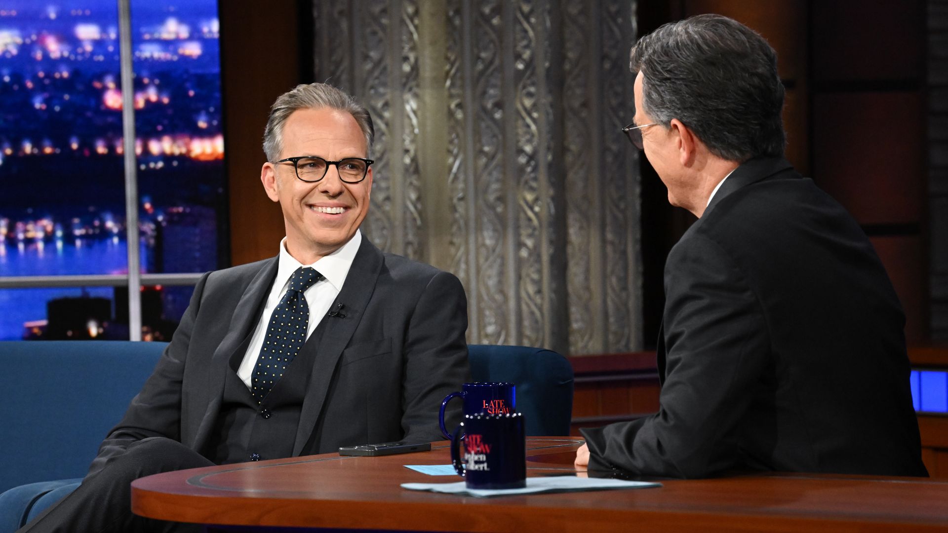 Jake Tapper in black suit and glasses smiling, in conversation with Stephen Colbert, seated at desk