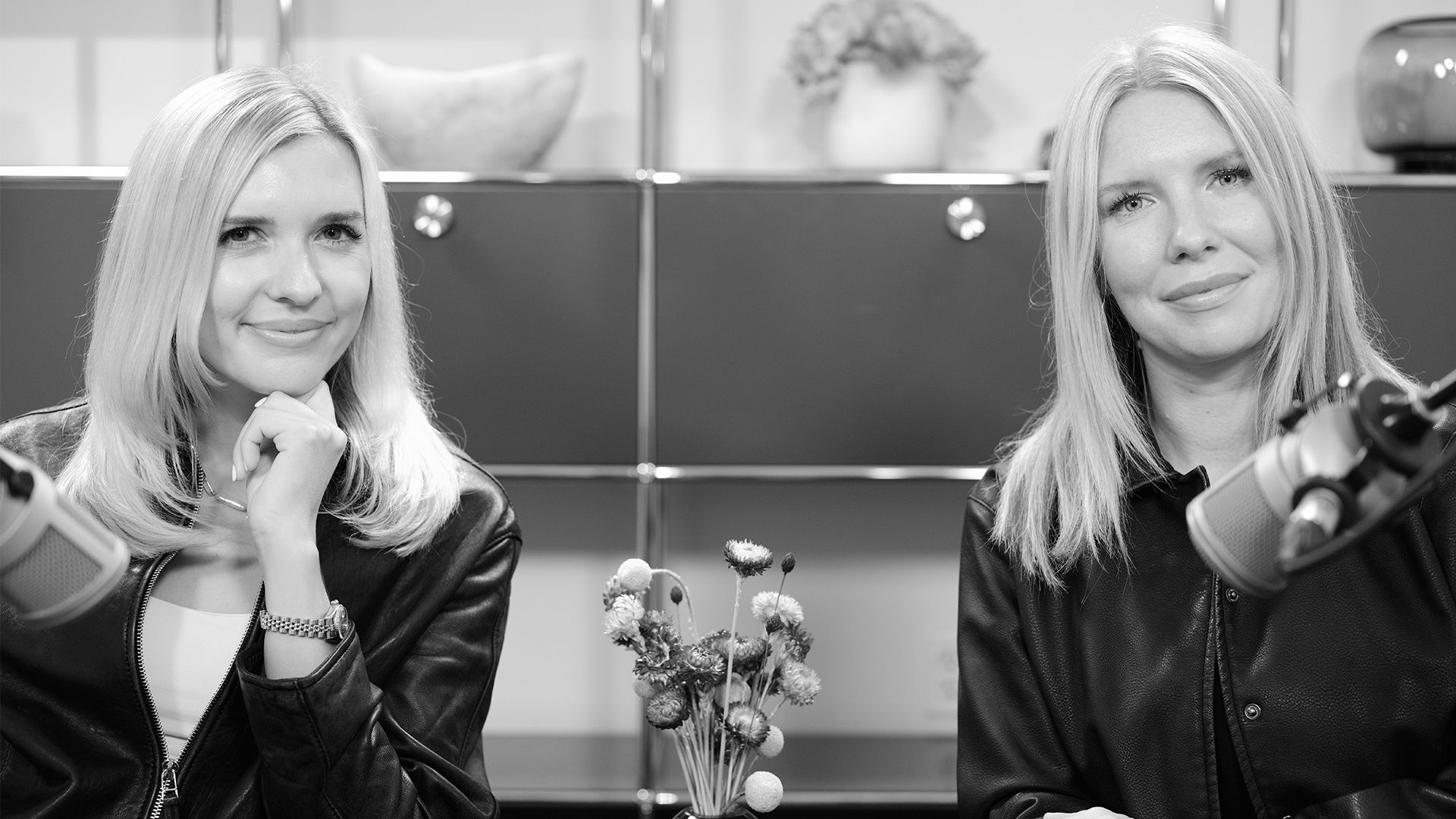 Two blonde women in black leather jackets sit in front of microphones with a small flower arrangement between them in a podcast or interview setting.