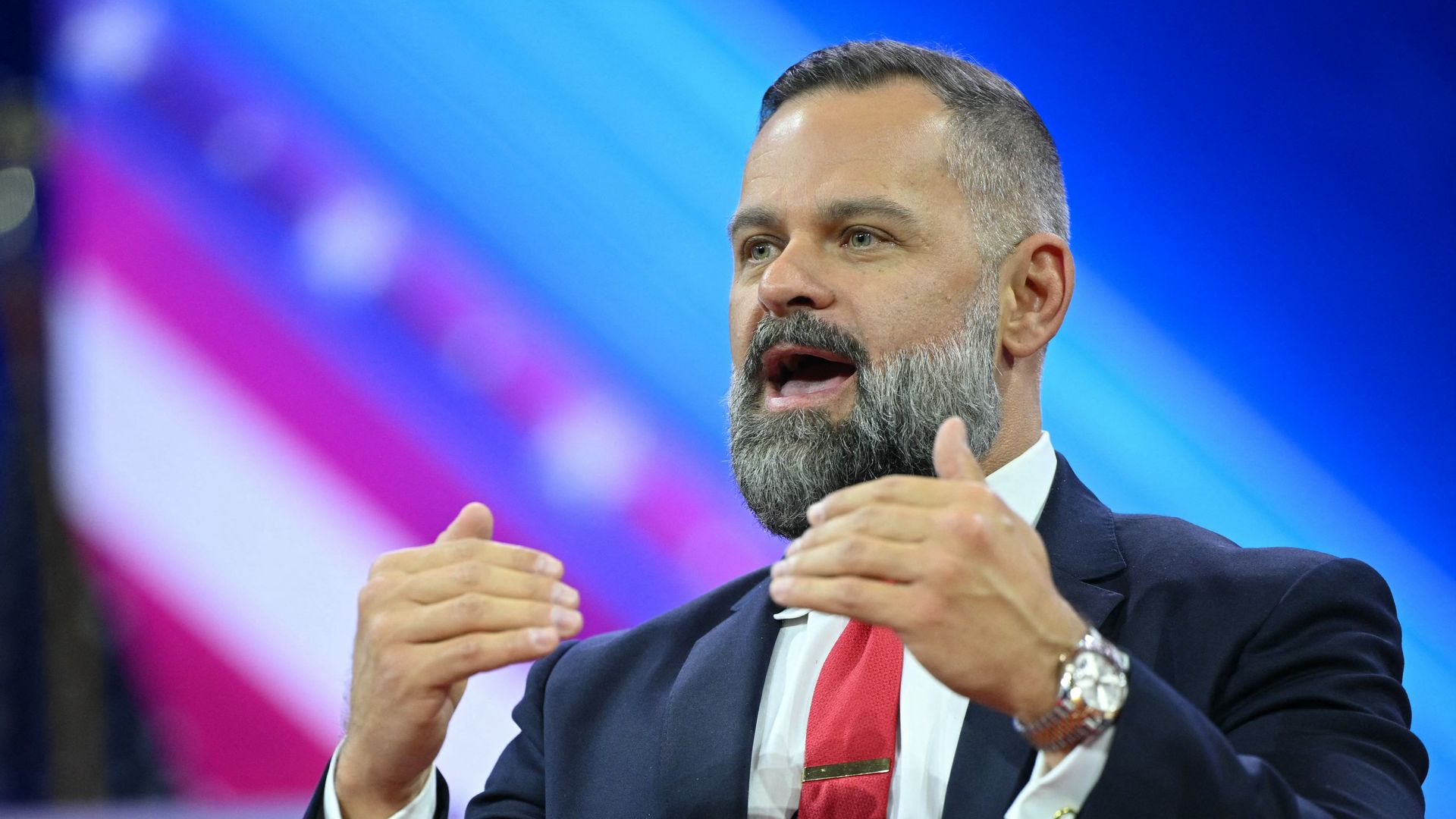 Cory Mills, wearing a blue suit and speaking while standing in front of a red, white and blue backdrop.
