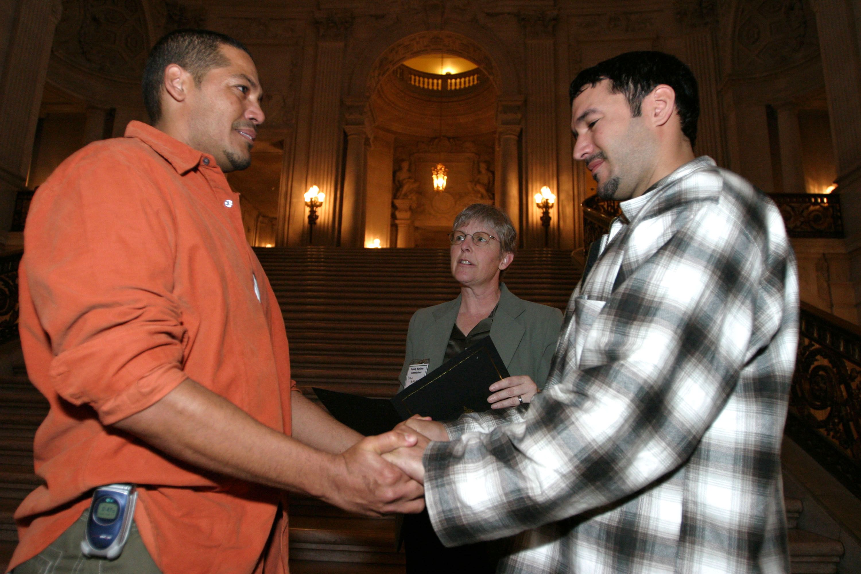 Photo of two men holding hands as a marriage officiant speaks behind them