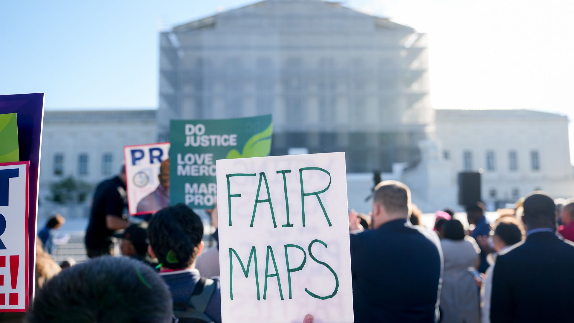 Demonstrators gather in front of the United States Supreme Court, which is covered in scaffolding. A handwritten sign in the foreground reads "fair maps" and several others hold signs in the background. 