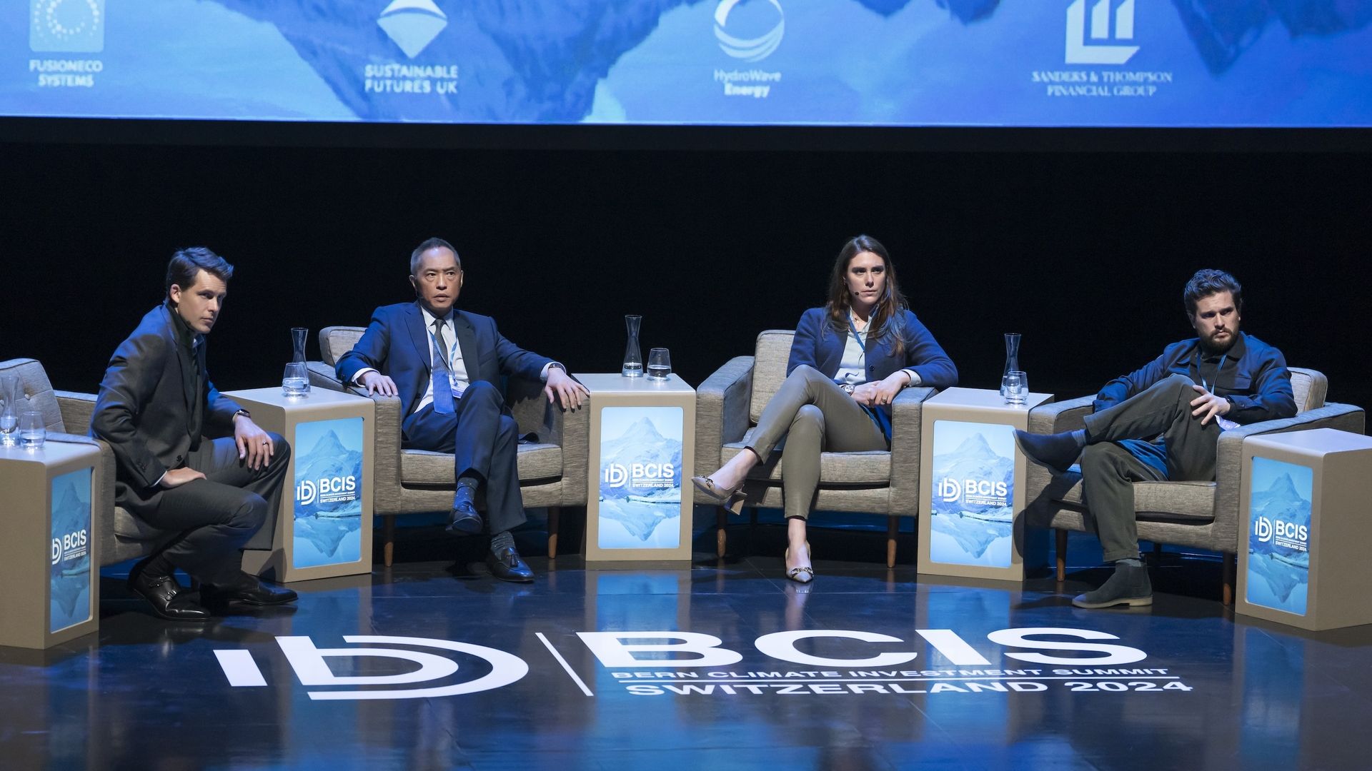 Still image from HBO's "Industry" Season 3, showing characters sitting on panel at climate conference stage.