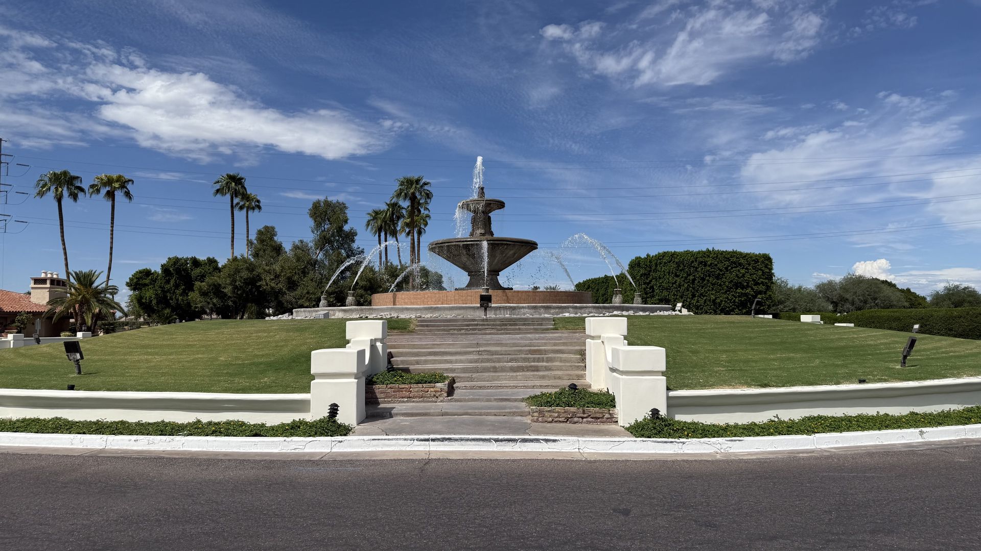 Wide view of a public plaza with a multi-tier stone fountain at center, water jets arching outward. Palm trees, green lawn, hedges, blue sky with wispy clouds, and a road in the foreground.