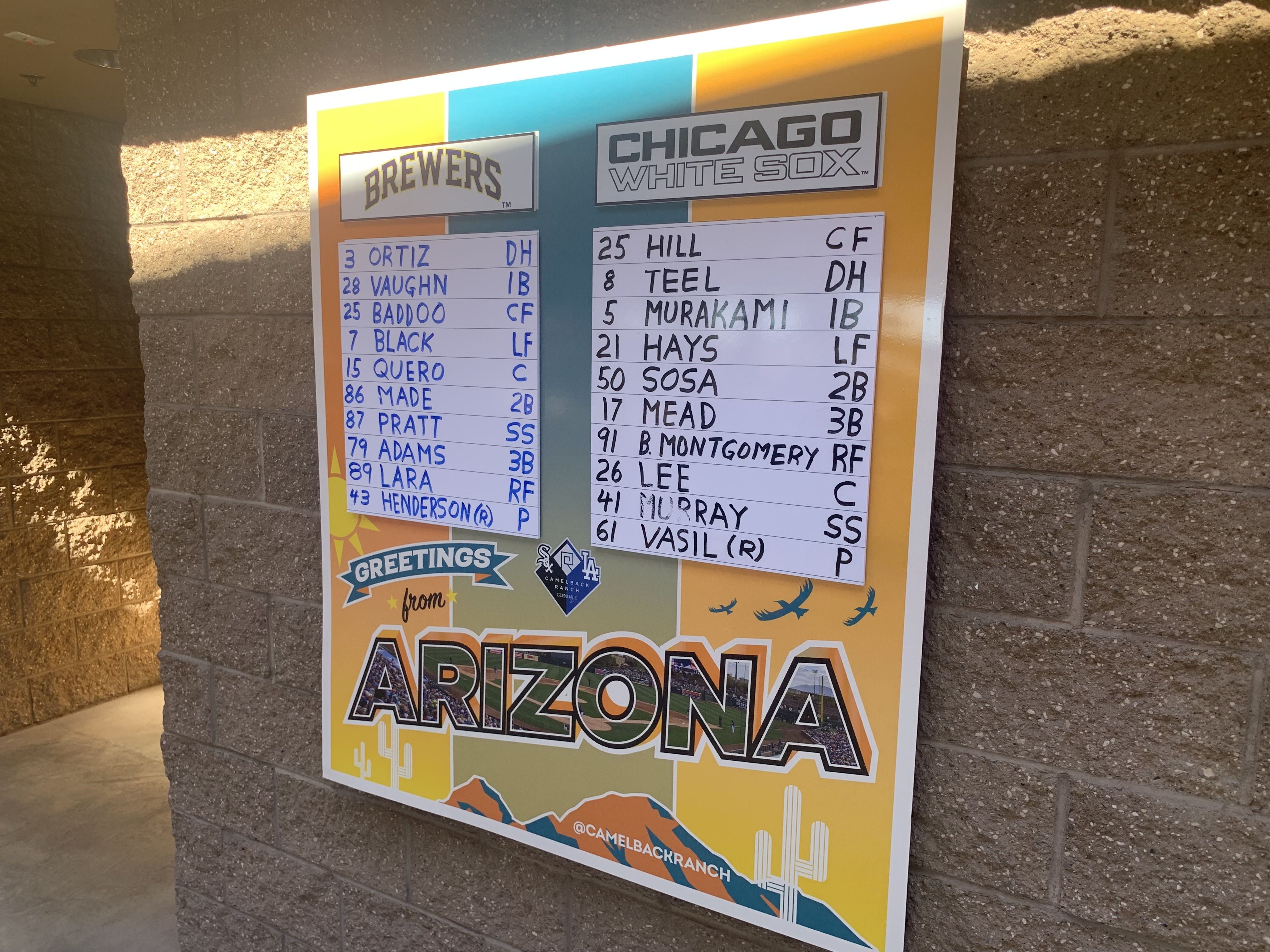 Baseball lineup board showing Brewers and Chicago White Sox players against a yellow and orange Arizona-themed background with cactus and mountains, mounted on a brick wall.