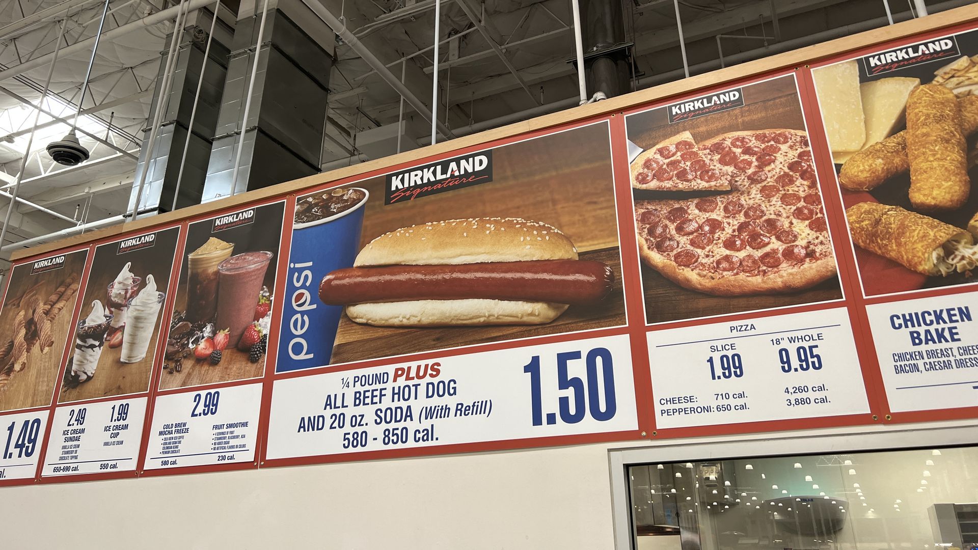 Industrial ceiling with exposed beams and ducts above a strip of large food posters displaying a hot dog, pepperoni pizza, and breaded sticks; Kirkland branding visible on the panels.