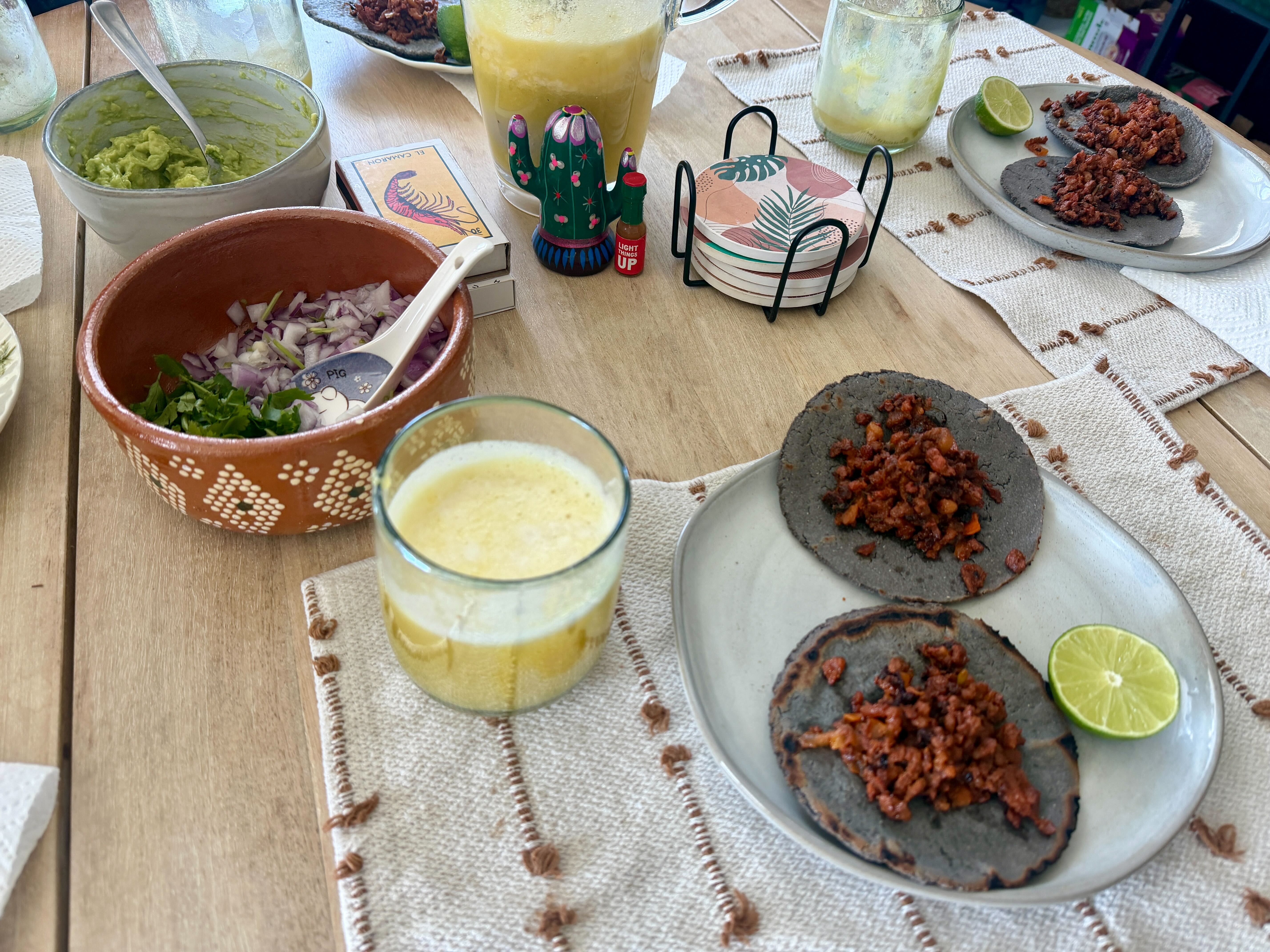 A wooden table with blue corn tostadas topped with seasoned meat and lime wedges, a plate of guacamole, chopped onions in a terracotta bowl, a cactus salt shaker, decorative plates, and yellow drinks.