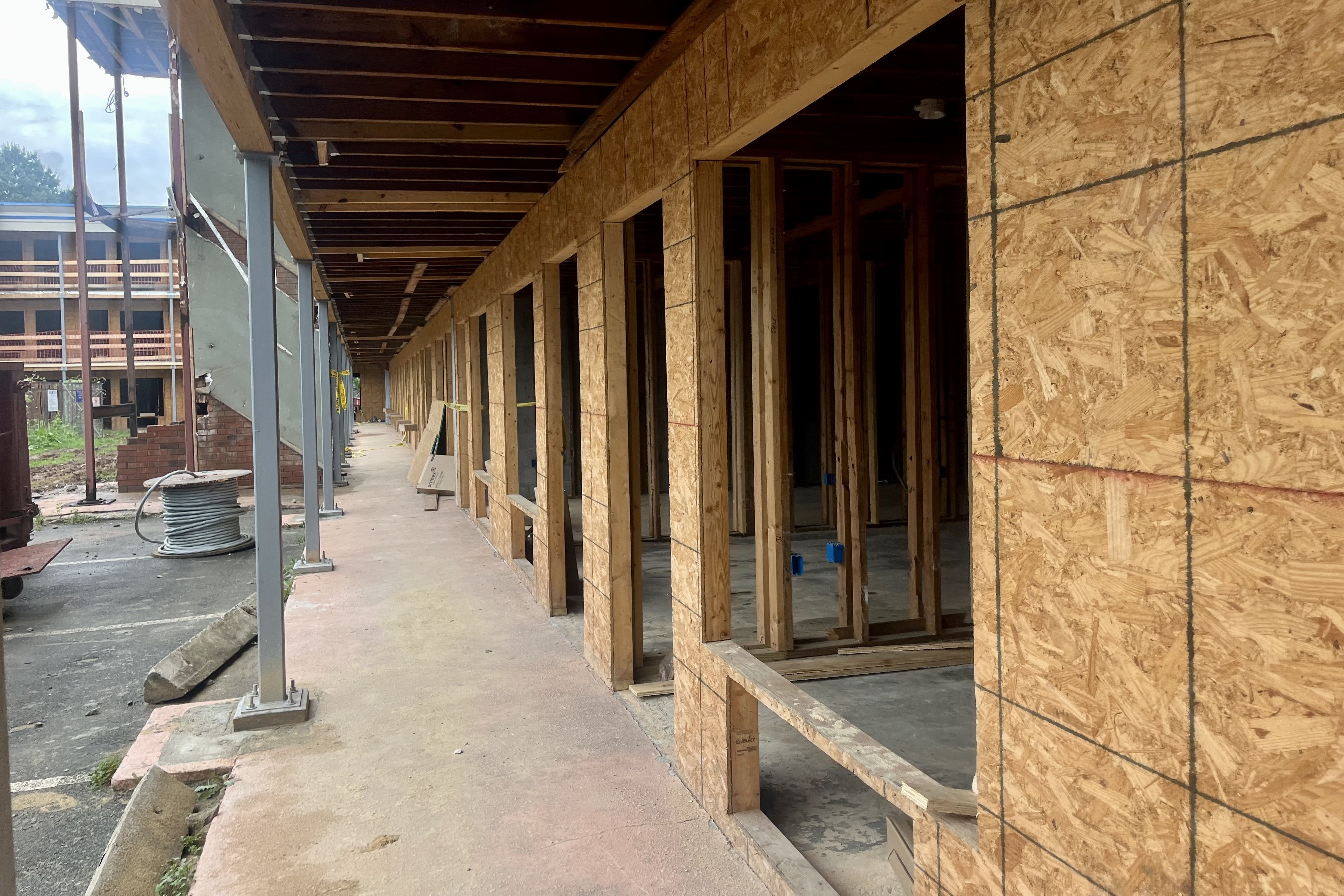 Long outdoor corridor of a building under construction with wooden framing and plywood walls, and an unfinished staircase with exposed steel beams on the left.