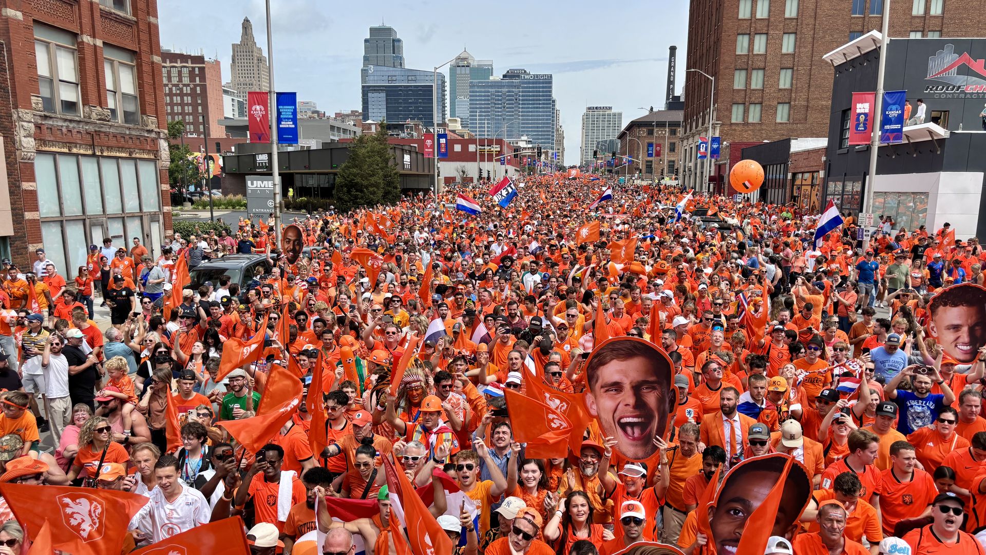 Thousands of people in orange shirts fill a city street for a parade, waving orange and national flags as tall buildings line the background with banners and storefronts.