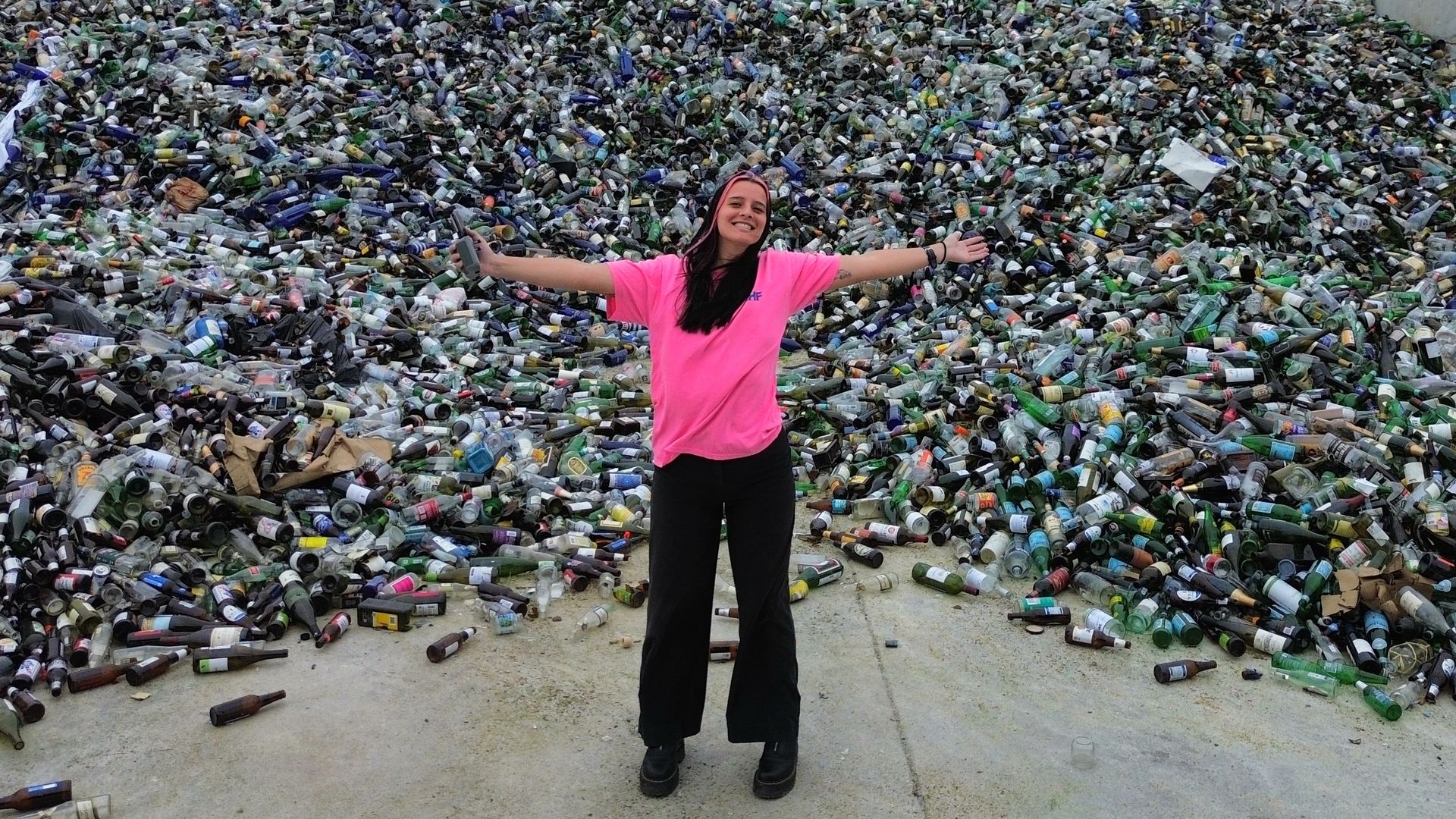 Smiling girl in a neon pink shirt stands with arms outstretched before a massive pile of discarded glass bottles inside concrete barriers, beneath a cloudy sky.