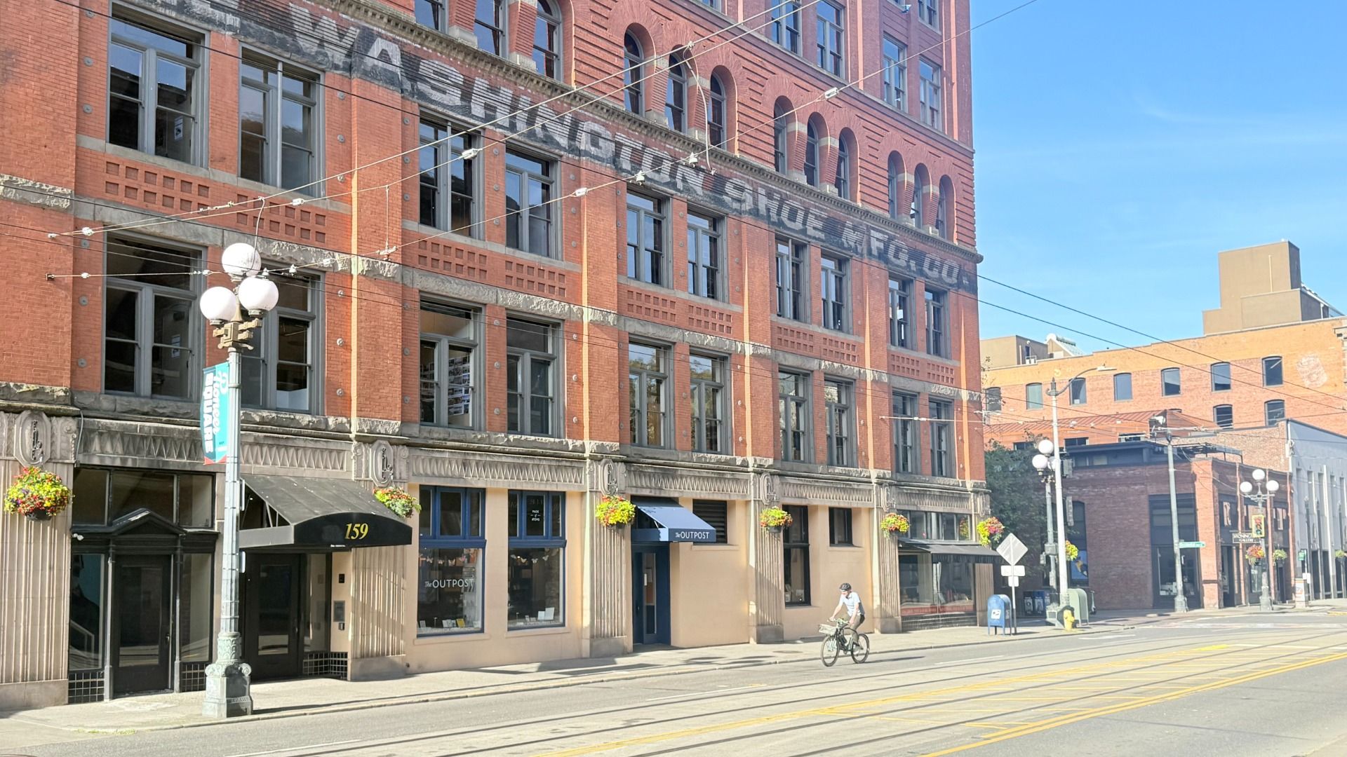Sunny city street with a red brick, multi-story building featuring arched windows and a faded banner along the facade. A cyclist rides by shops with hanging planters and string lights overhead.