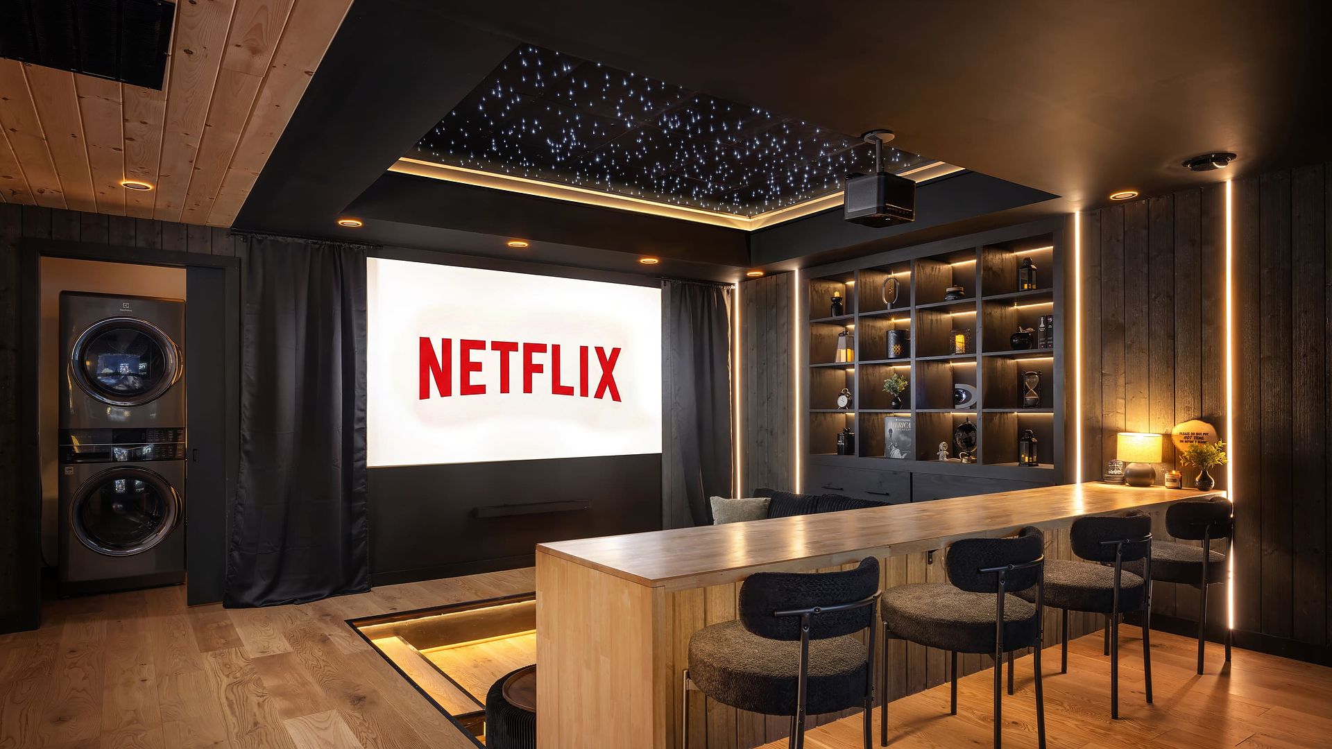 Cozy home theater with a large "Netflix" projection, a wooden bar and stools, warm LED lighting, and built-in shelves. A starry ceiling, dark curtains, and a laundry stack on the left.