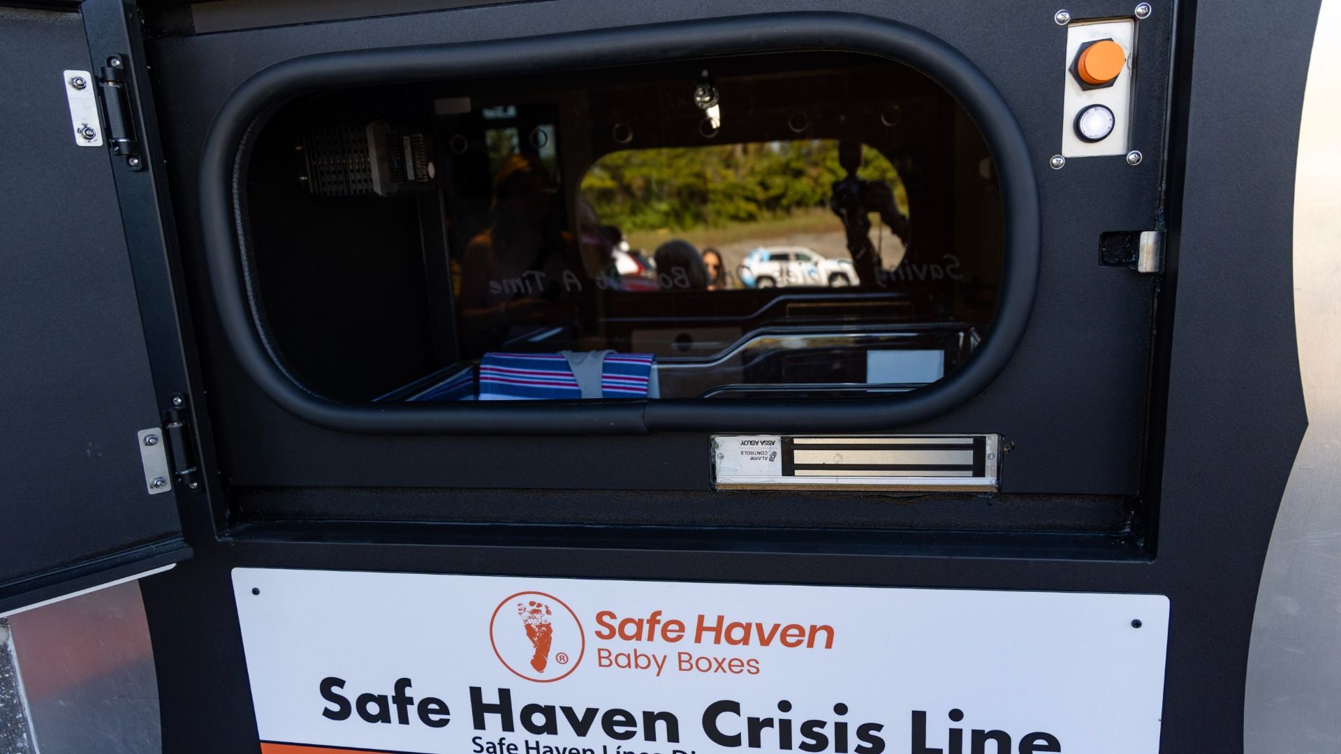 Close-up of a Safe Haven Baby Boxes unit: dark metal exterior, rounded viewing window, orange emergency button and white indicator light; inside a striped cloth rests on a padded surface, with reflections of people and cars outside.