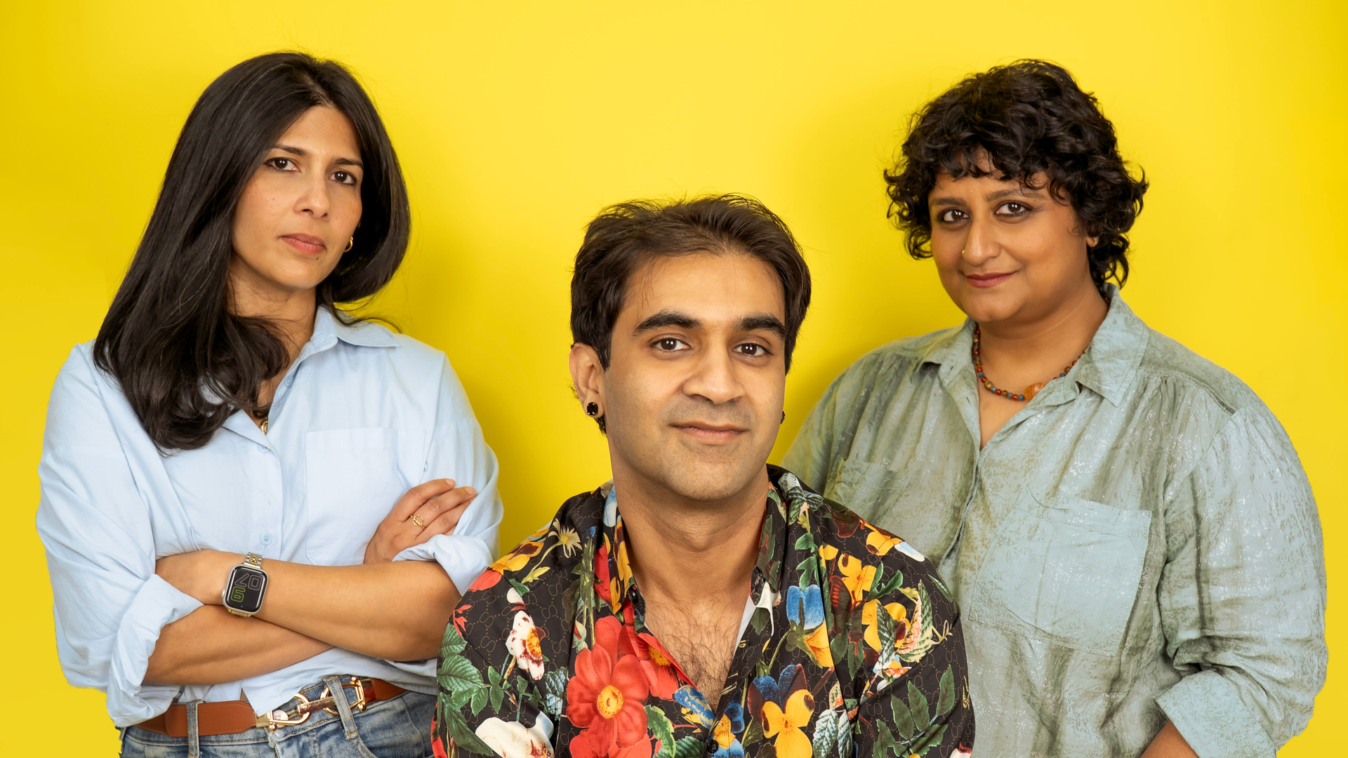 Three people pose against a bright yellow backdrop: left woman in light blue shirt, center man in tropical floral shirt, right woman in light green shirt.