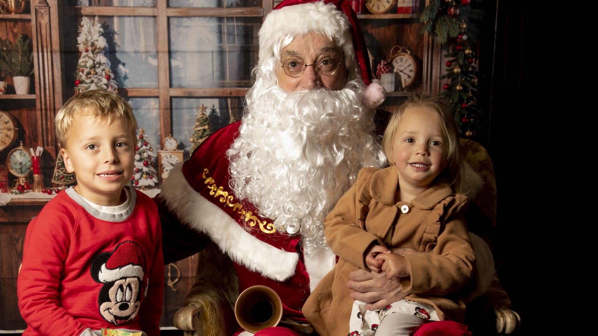 Two small kids with one sitting on Santa's lap and the other standing nearby
