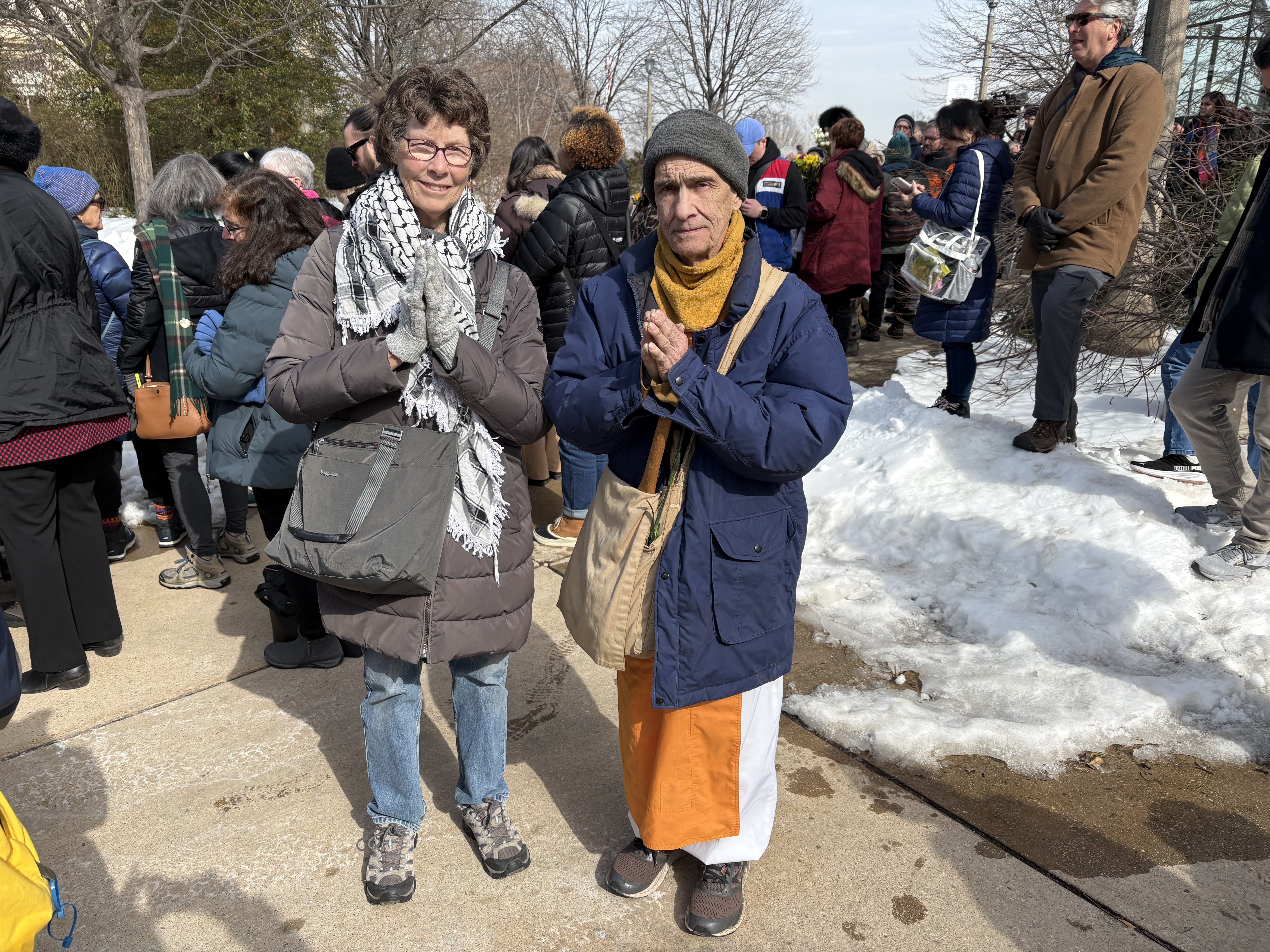Two people standing outdoors in winter clothing, one in a blue coat and gray hat, the other in a gray coat and scarf, both with hands pressed together in a prayer-like gesture, snow on the ground.
