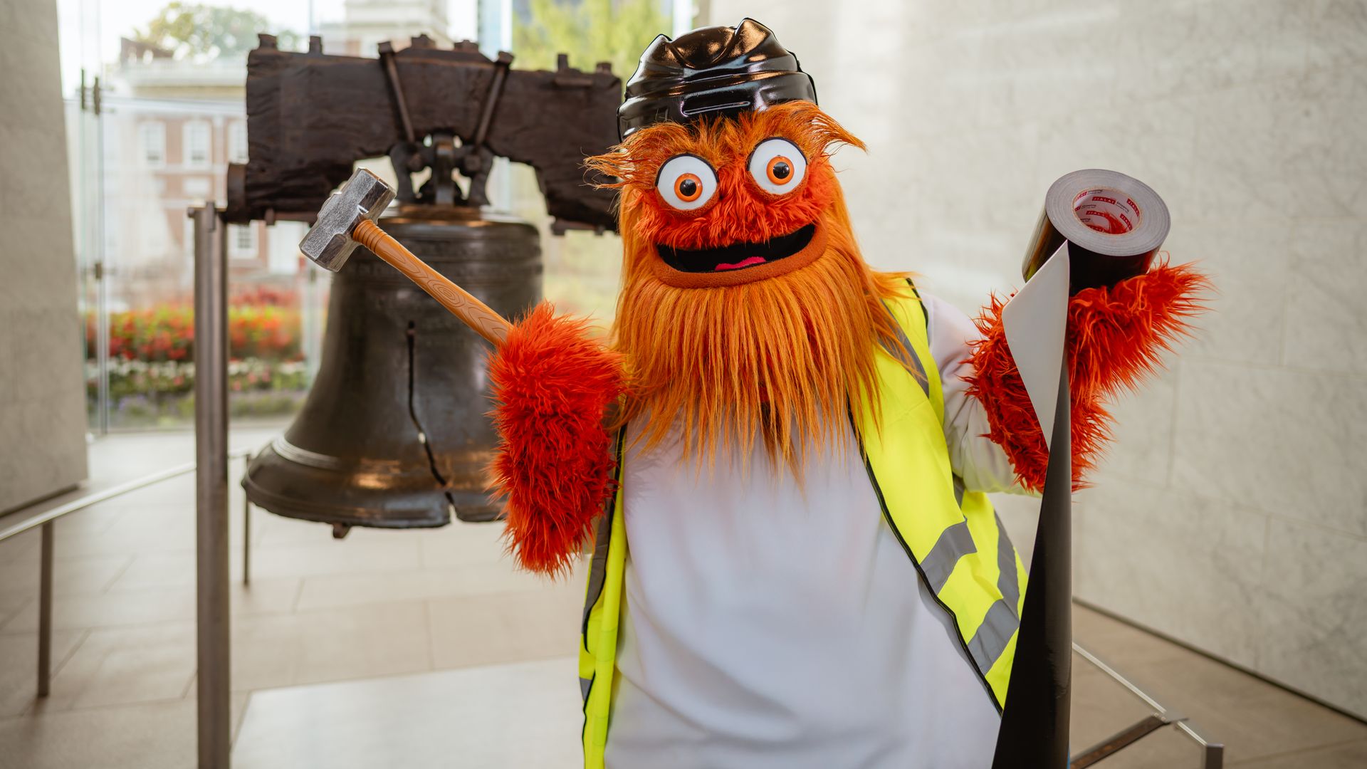 Flyers mascot Gritty wearing a black helmet and yellow safety vest, holding a hammer and tape, standing in front of Philly's Liberty Bell.