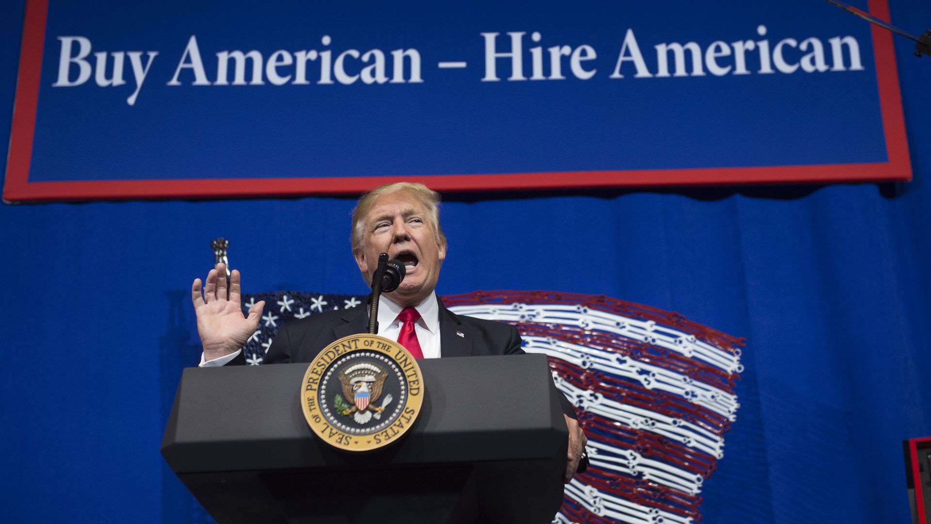 Donald Trump speaking at an event with a sign behind him that says "Buy American - Hire American"