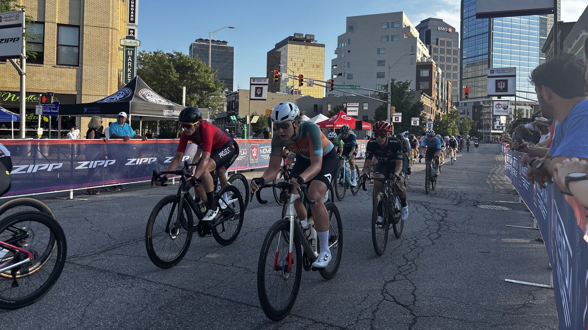 Group of cyclists racing through a city street; barriers read 'ZIPP,' tall buildings line the road, clear blue sky above, and spectators along the barriers.