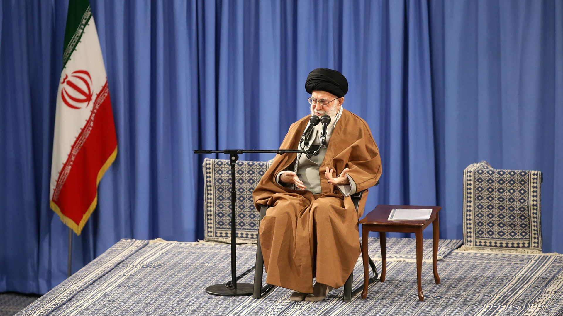 Khamenei seated in a chair on stage, next to the Iranian flag