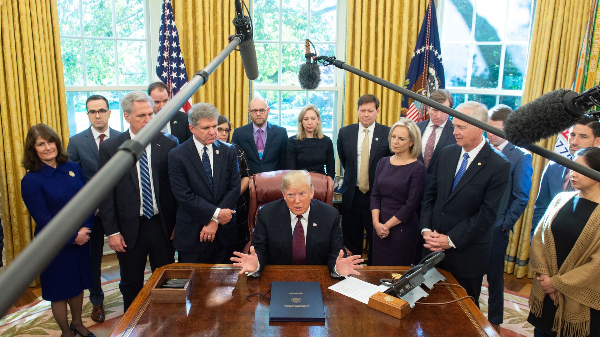 Photo of Donald Trump sitting in the Oval Office with officials standing around him and two mikes hovering over them
