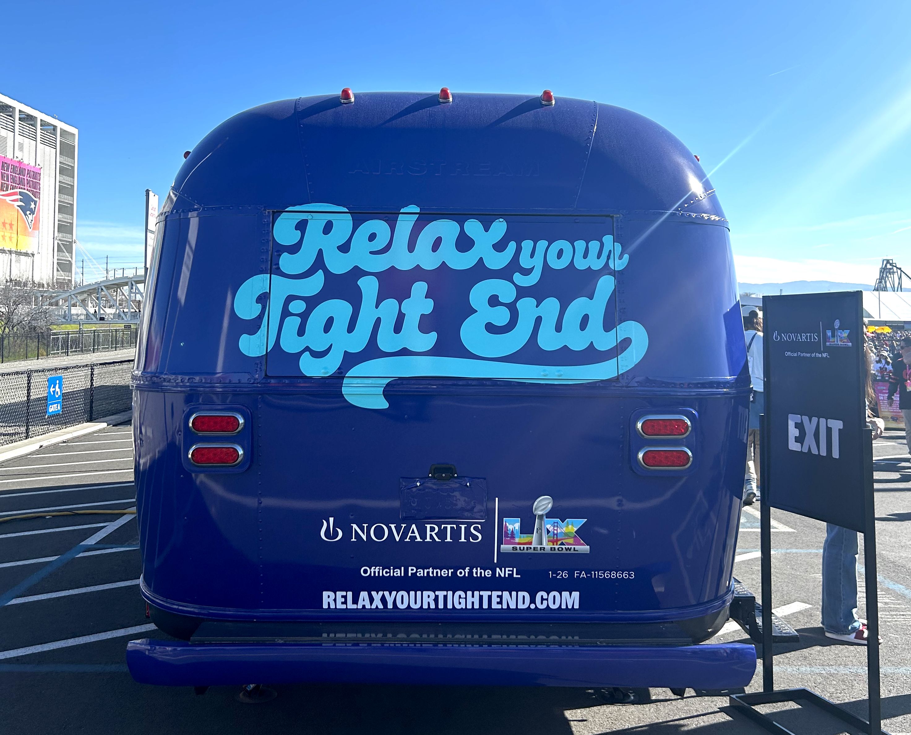 Back of a blue vehicle with large light blue text "Relax your Tight End" and Novartis NFL partnership logos, with a sign next to it reading "EXIT" on a sunny day.