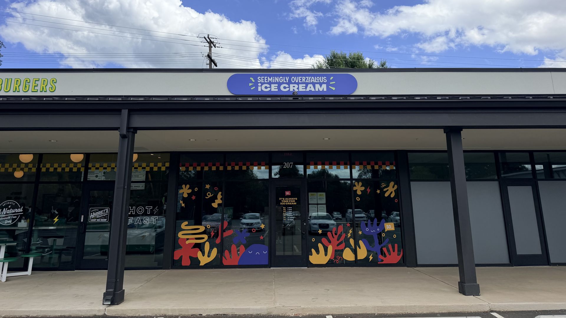 Wide storefront under a blue sky with white clouds. A blue oval sign reads "SEEMINGLY OVERZEALOUS ICE CREAM" above the entrance; colorful handprint window art decorates the glass, with a partial "BURGERS" sign to the left.