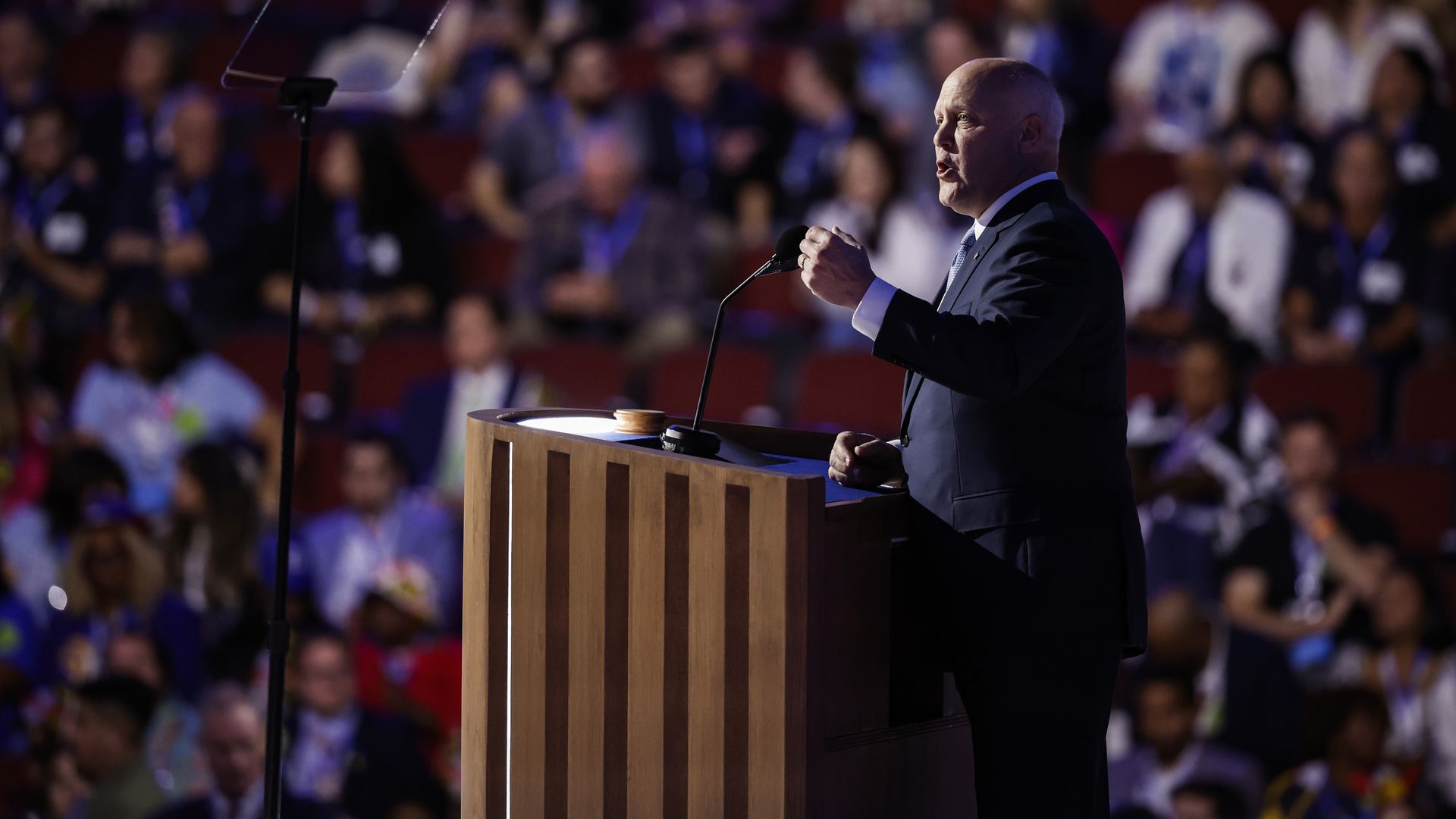 Mitch Landrieu, wearing a suit, speaks at a podium in profile. A crowd is in stadium seating behind him.
