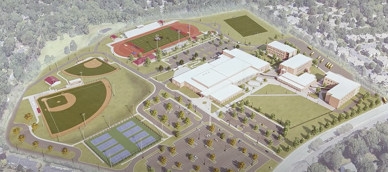 Aerial view of a large school campus with multiple buildings, a football field with running track, two baseball fields, tennis courts, a parking lot, and surrounding trees and houses.
