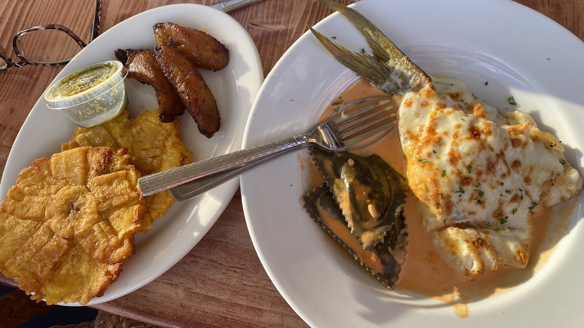A small white plates holds tostones and plantains. A bigger plate holds a fish covered in melted cheese.