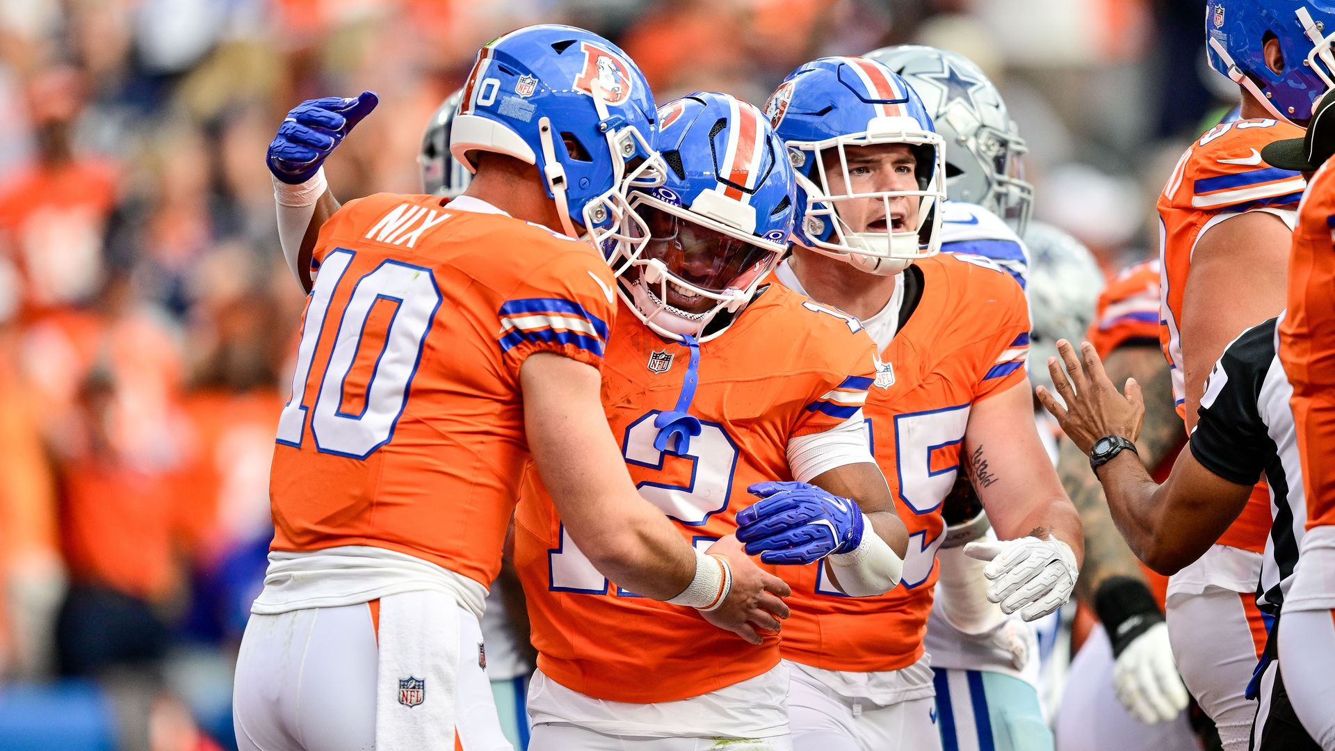 Men in blue football helmets and bright orange jerseys smile on and celebrate during a sunny day. 