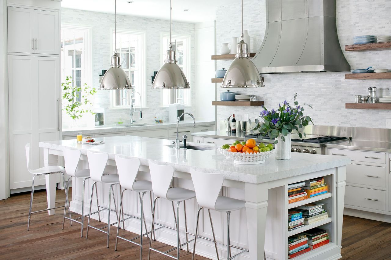 Bright white kitchen with a large island featuring six white stools, a fruit bowl, and a vase of purple flowers. Stainless steel range hood and three pendant lights hang overhead.