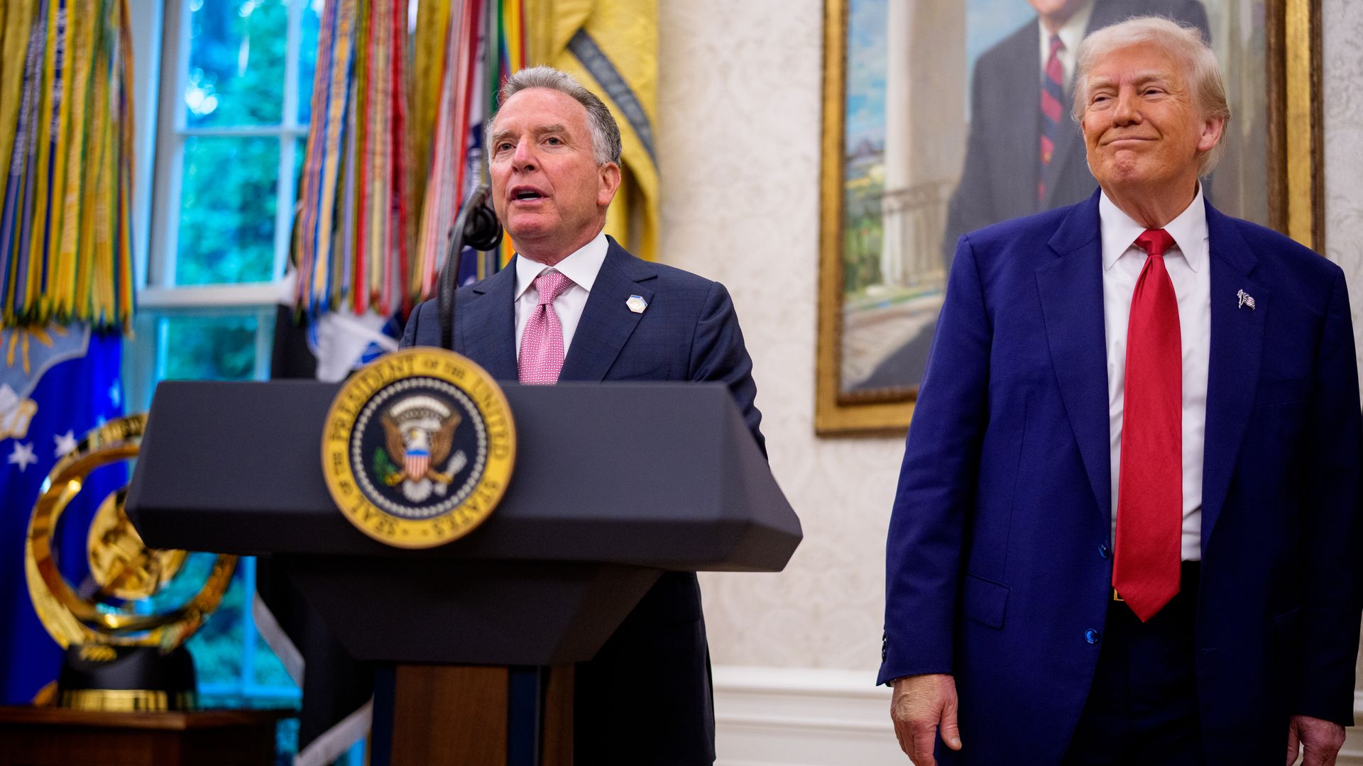 Man in dark suit with pink tie speaking at a lectern with the presidential seal, another man in a blue suit and red tie standing beside him in a room with military flags and a large portrait on the wall.