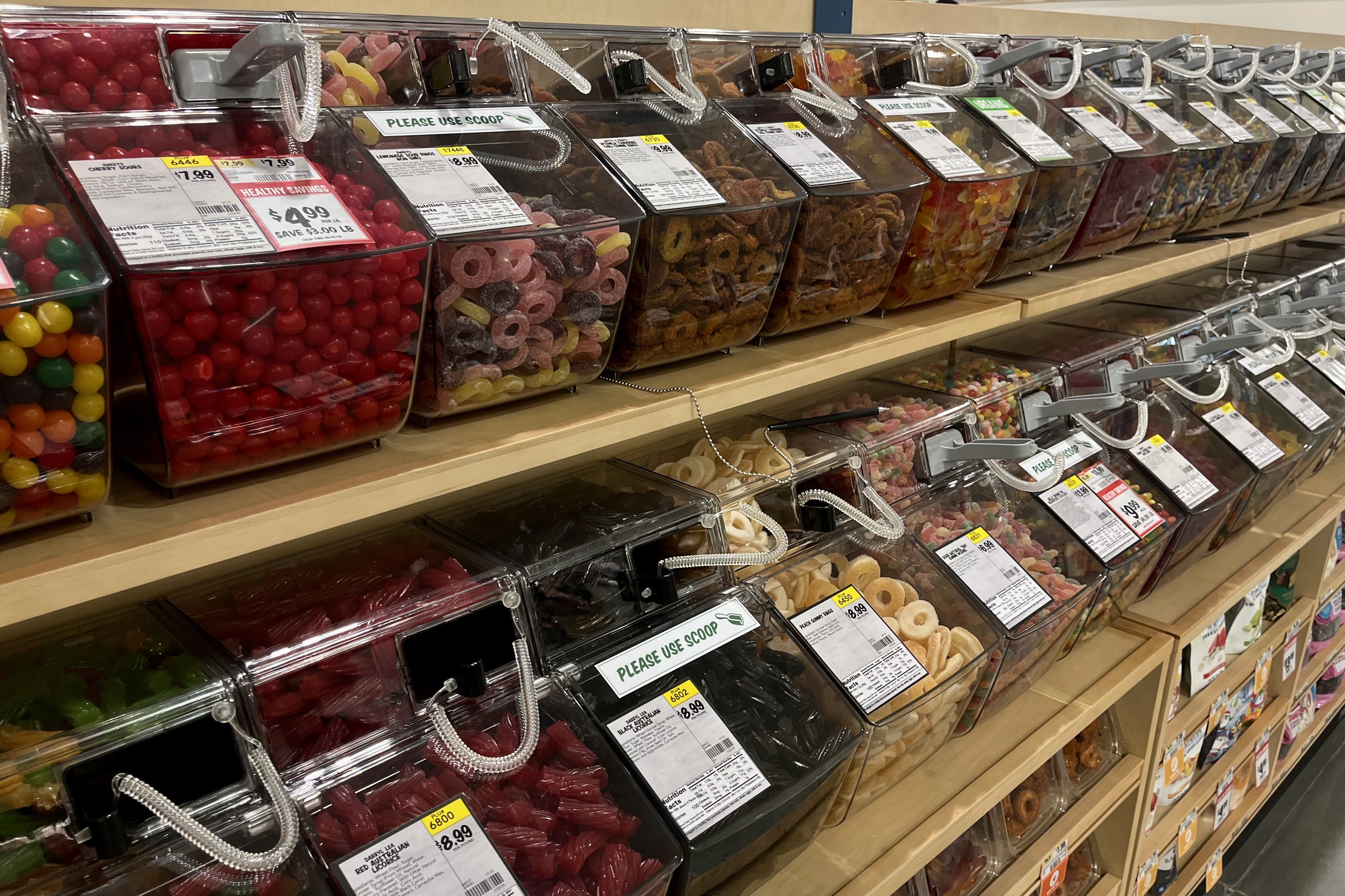 Two wooden shelves filled with clear plastic bins of assorted colorful candies including red cherry drops, gummy rings, licorice, and more, each labeled with price tags and scoop instructions.
